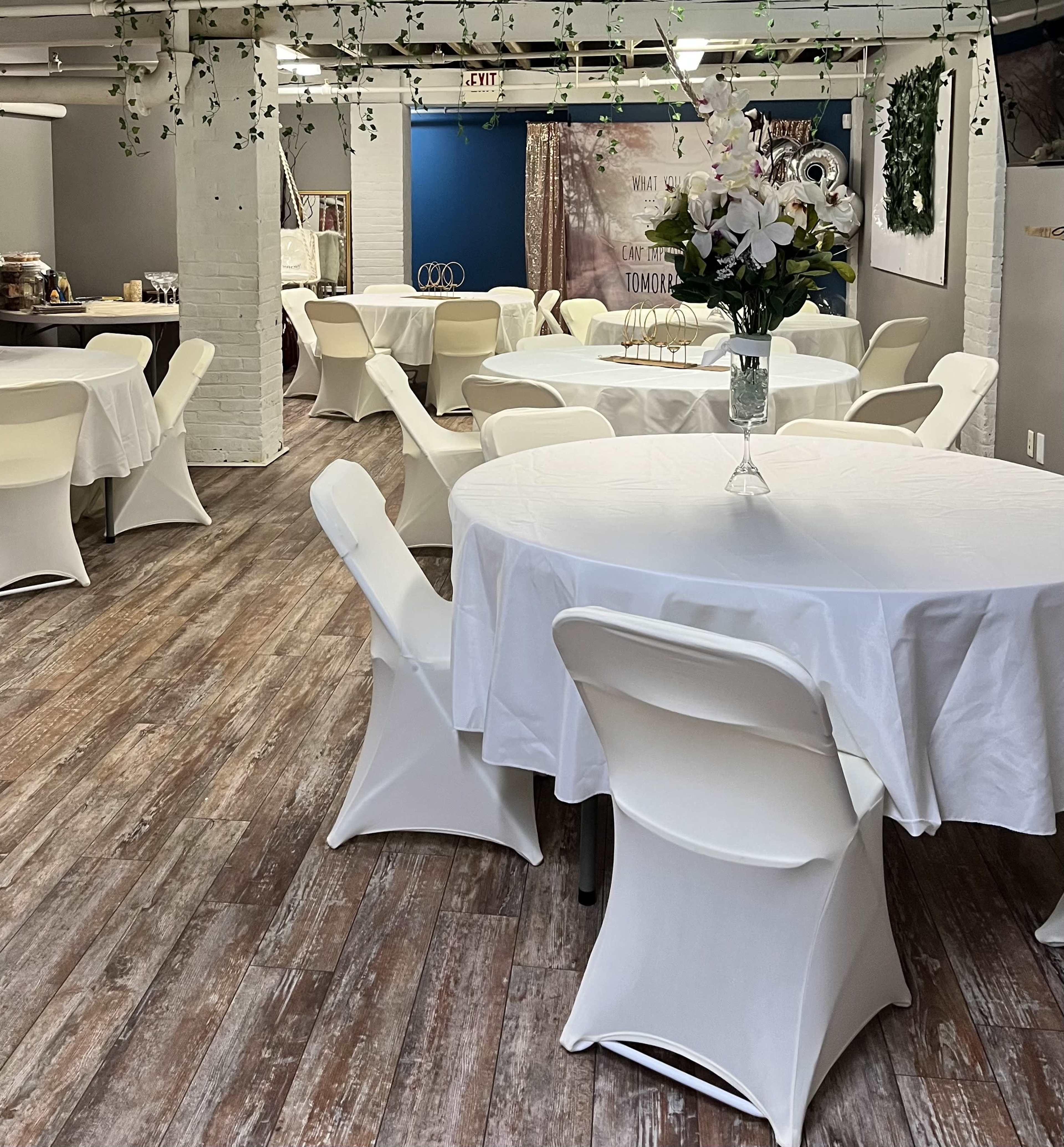 A spacious event room features white tables covered with tablecloths and surrounded by chairs, with decorative plants hanging from the ceiling.