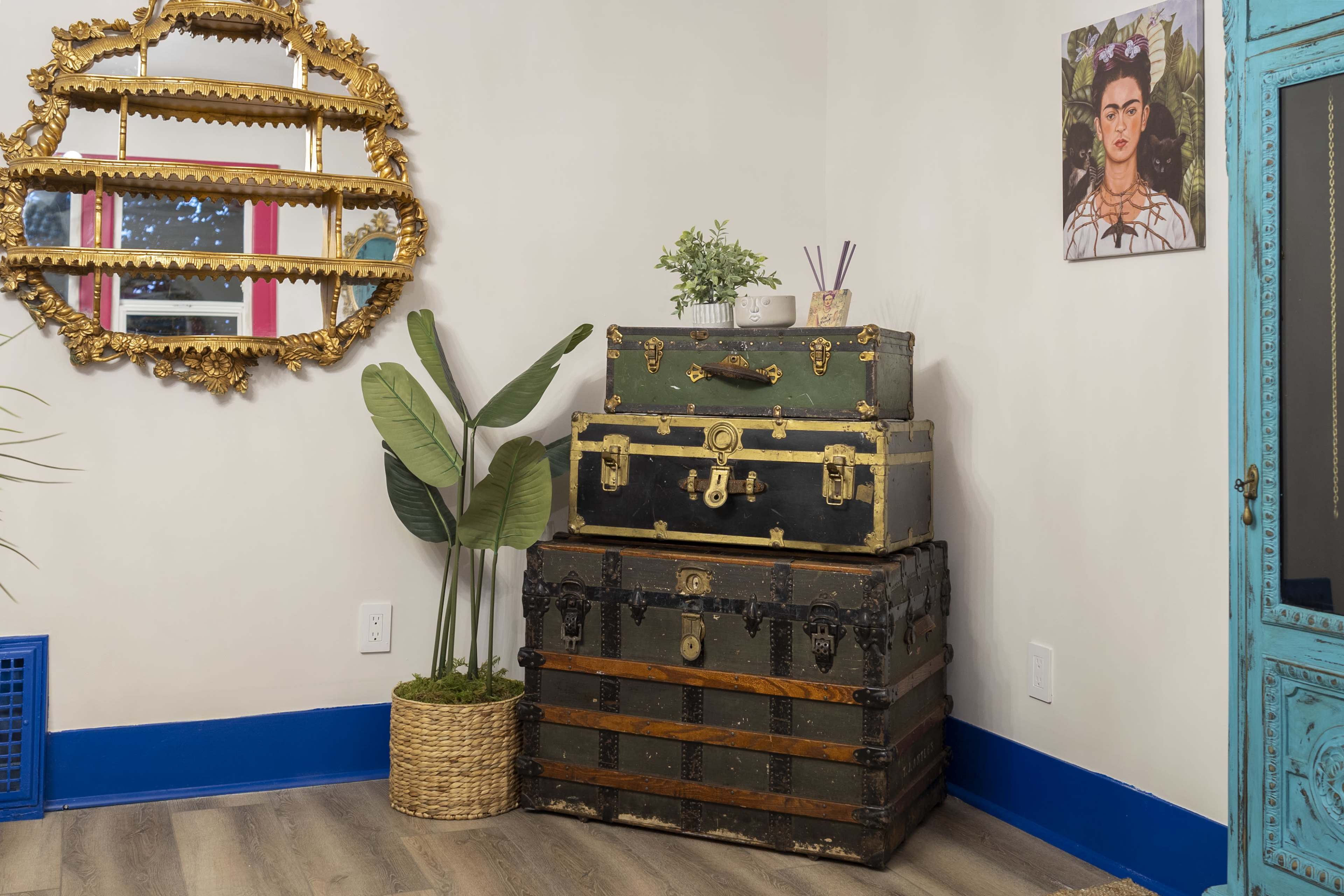 A stack of vintage trunks sits in a corner of a room, accompanied by a potted plant and a decorative mirror on the wall.