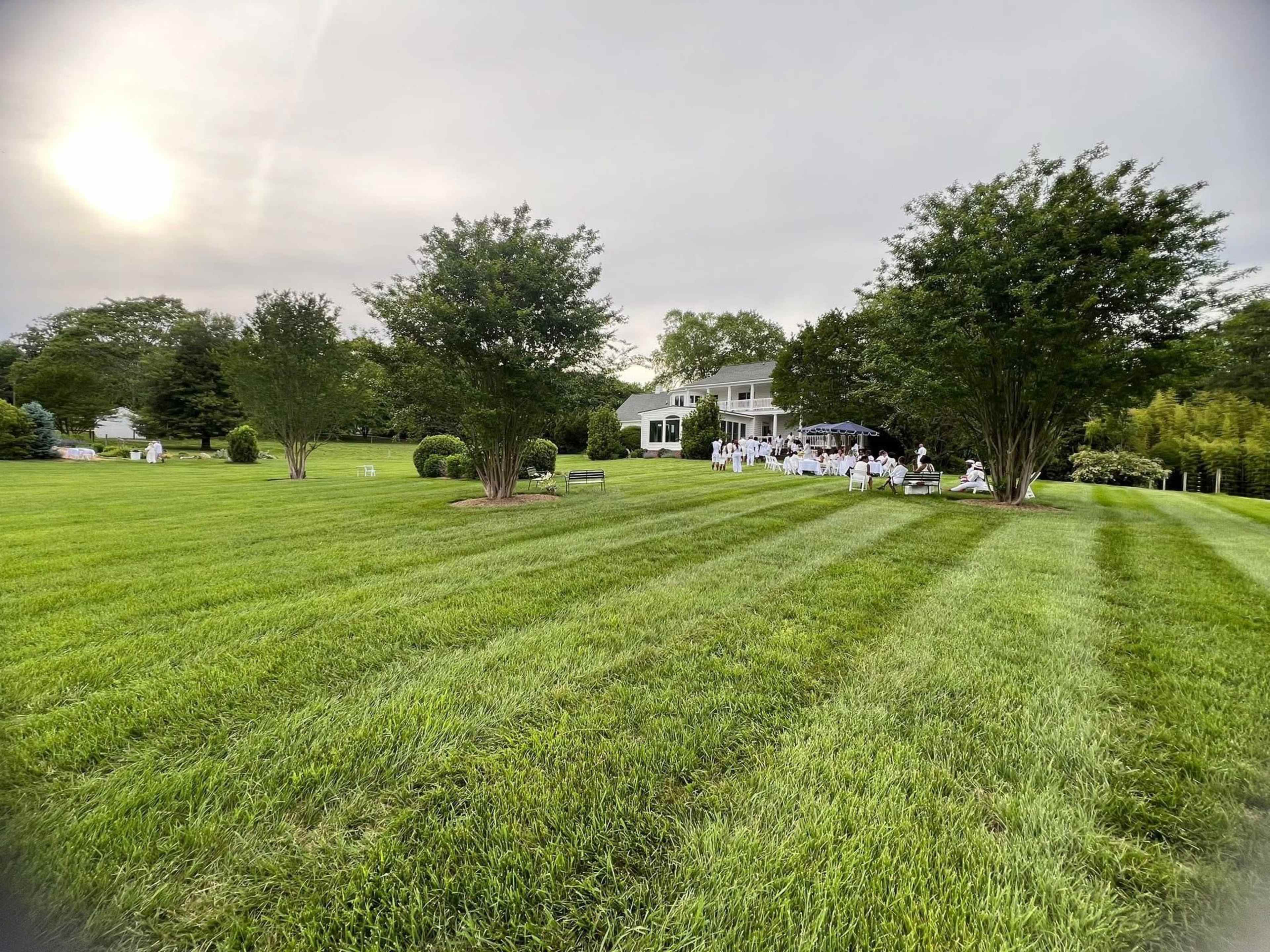 A white tent is set up in a grassy area with neatly trimmed lawn and trees, adjacent to a house.