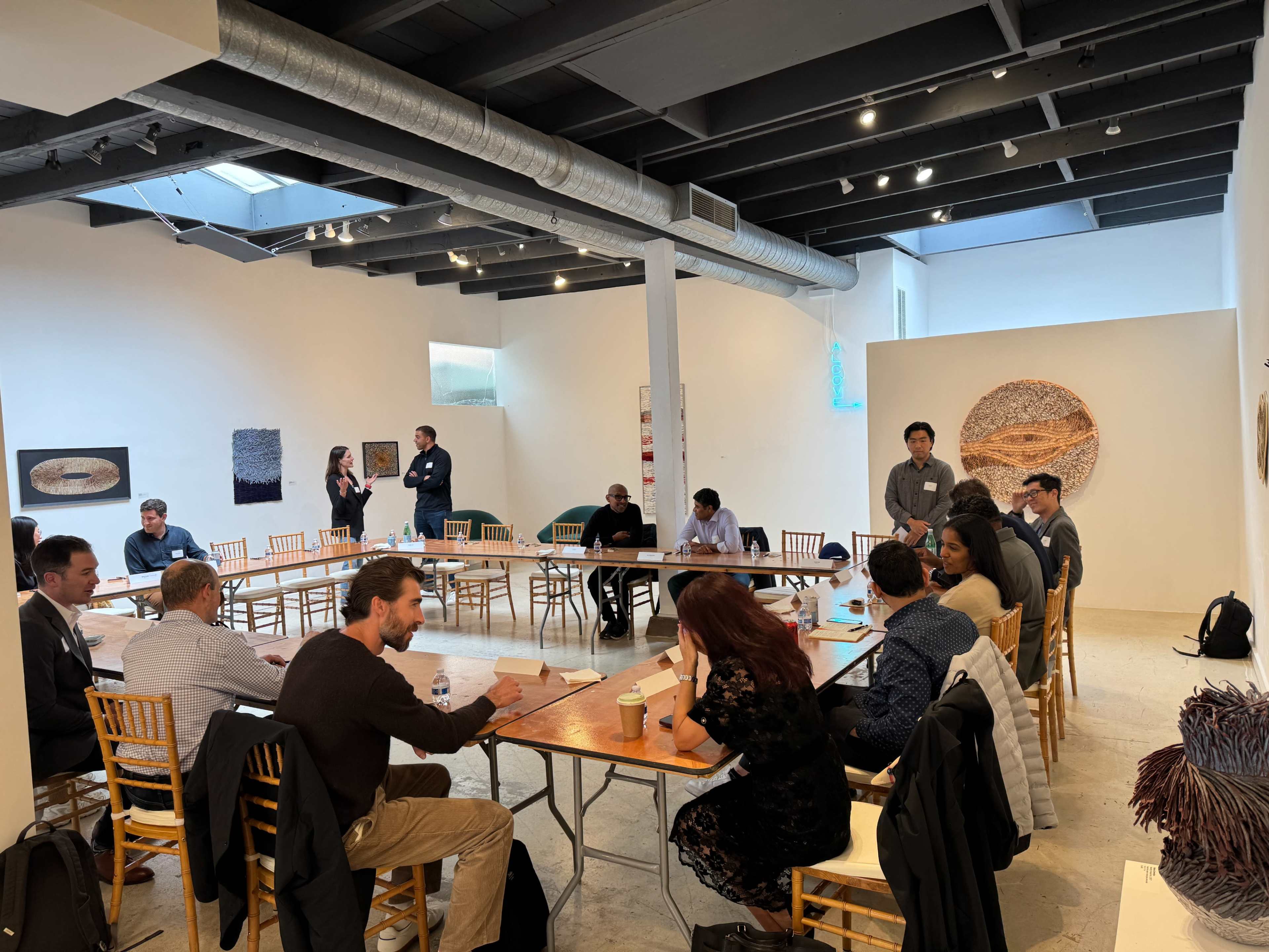 A group of individuals is gathered around several tables in an art gallery, engaging in discussions or activities.