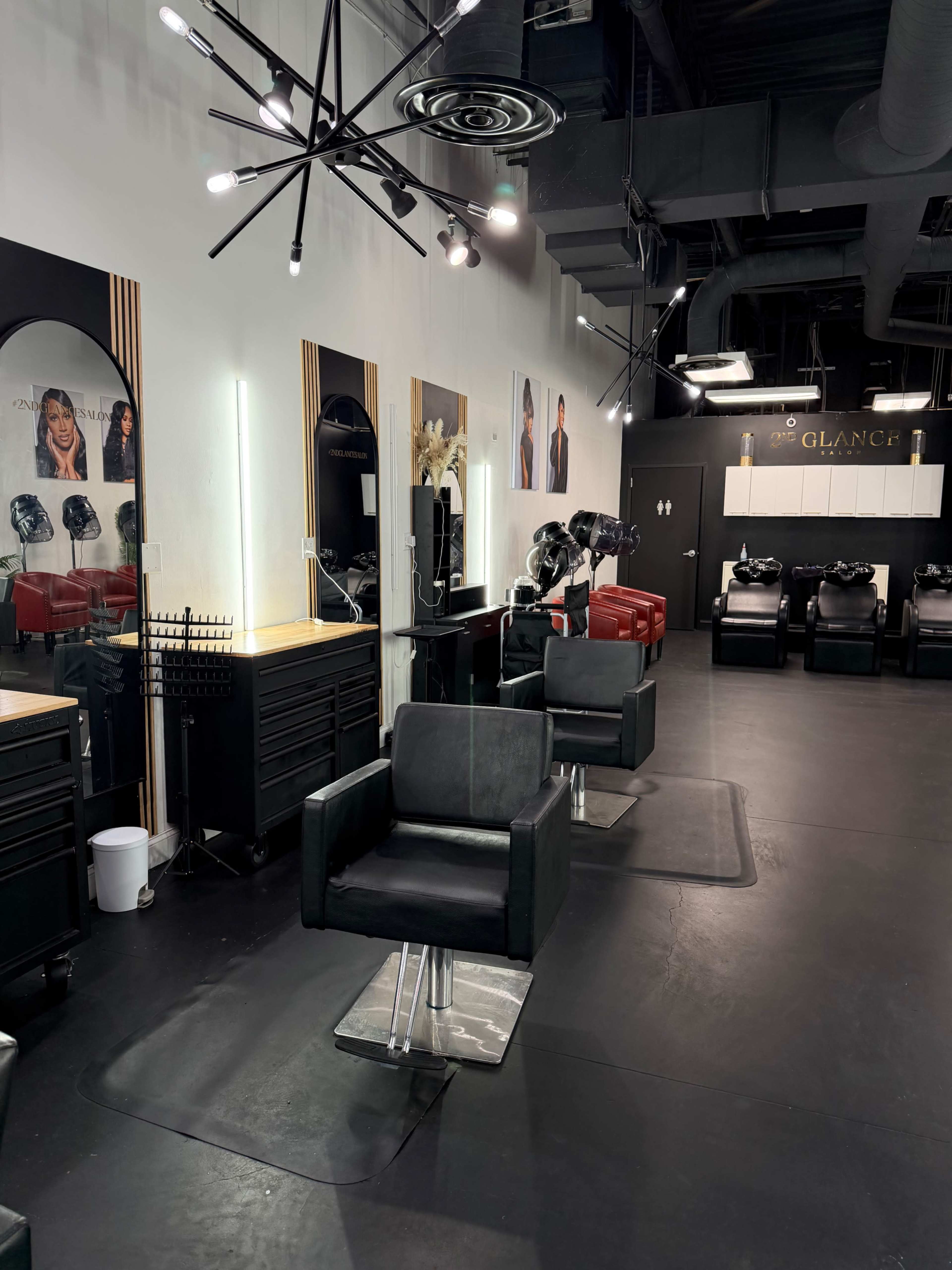 The image shows a modern hair salon with black chairs, mirrors, and stylists' stations, featuring a well-organized layout and a separate area for hair washing.