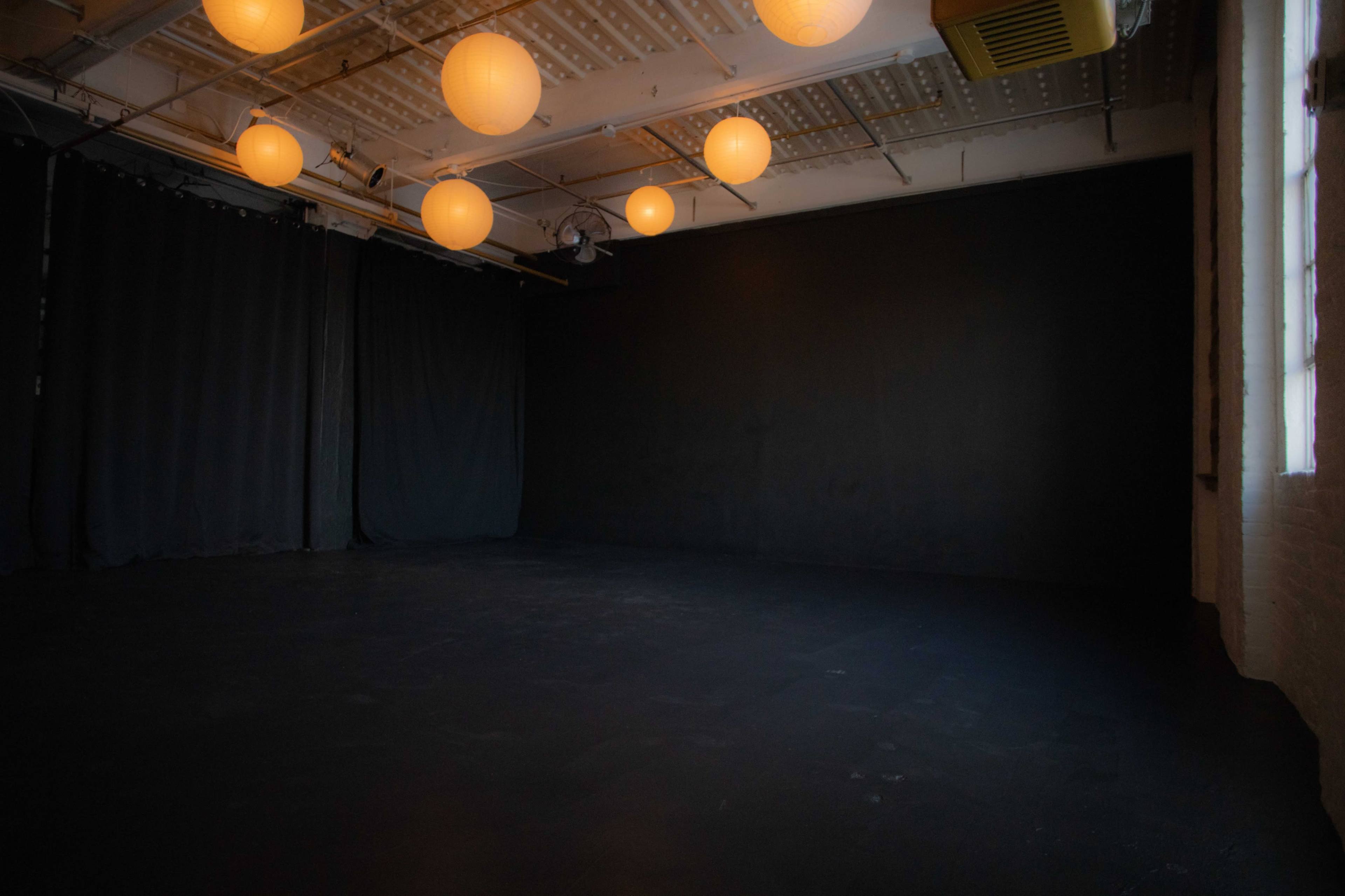 A spacious, dimly lit room with black walls and ceiling fixtures illuminating orange paper lanterns.