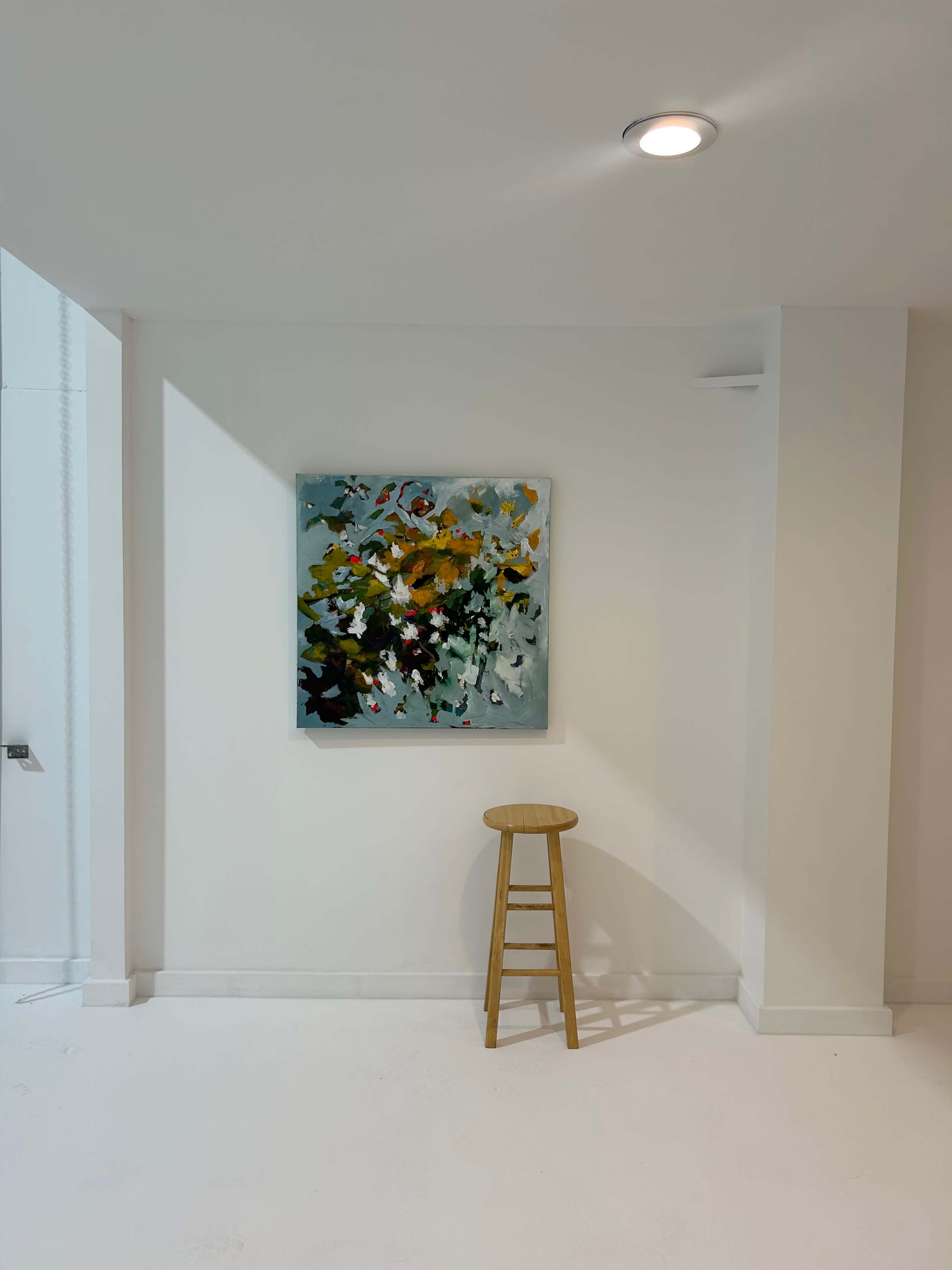 The image shows a minimalist interior space with a piece of abstract art hanging on a white wall beside a wooden stool.