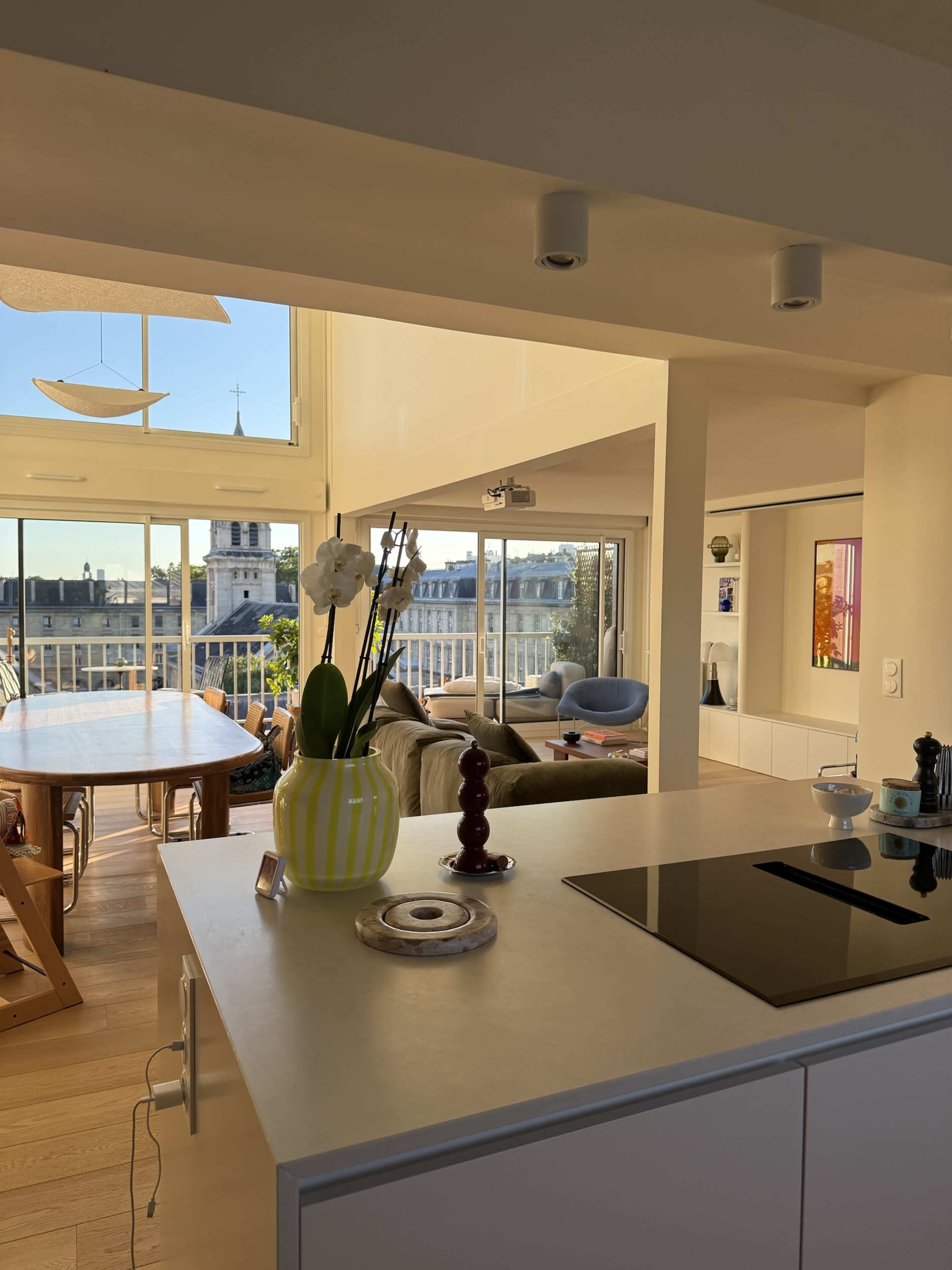 The image shows a bright, open-plan living space with a kitchen area leading to a dining table and a view of a cityscape through large windows.