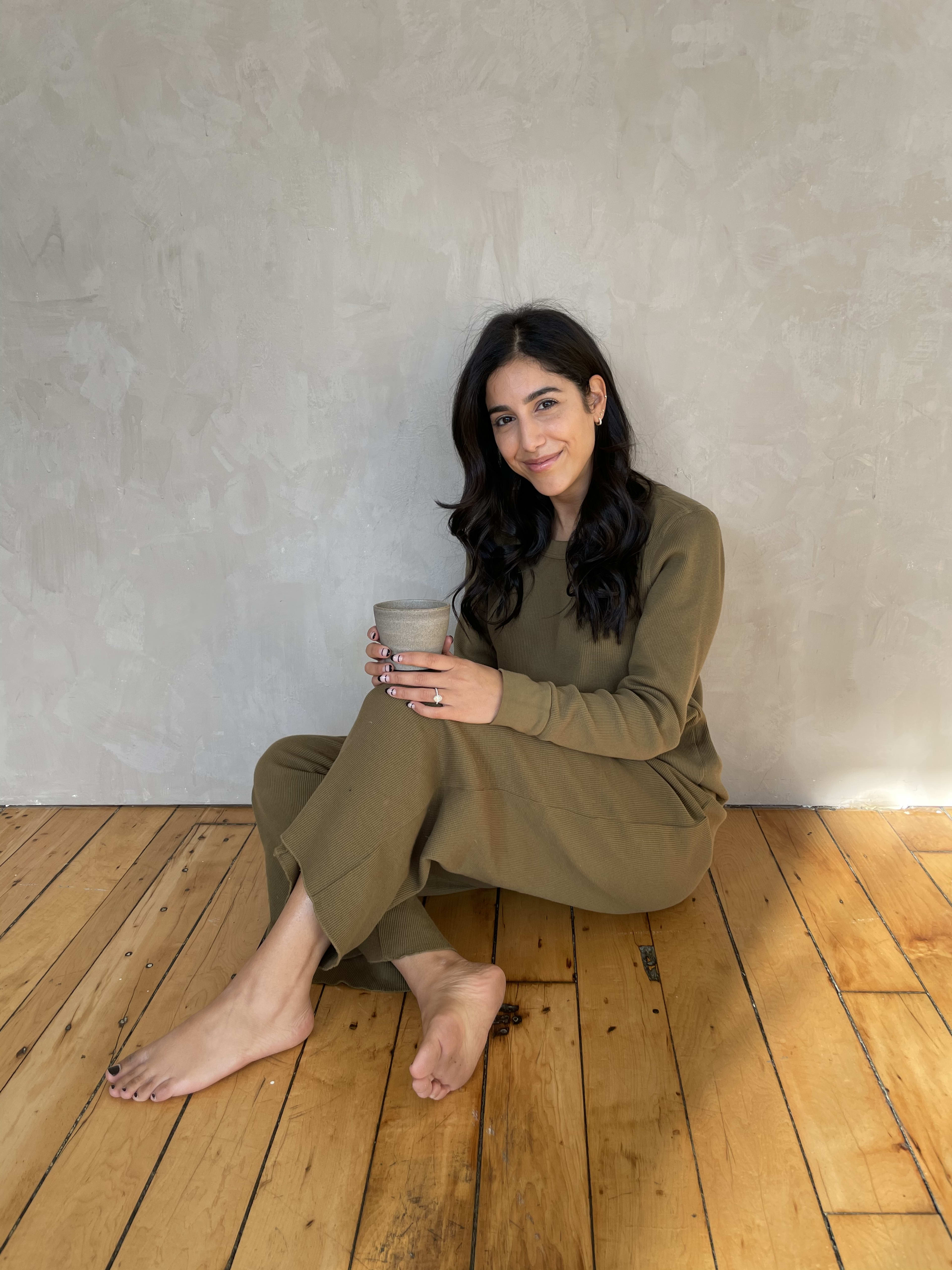 Une femme rustique tenant une tasse adossée à un mur gris lors d'un shooting photo.