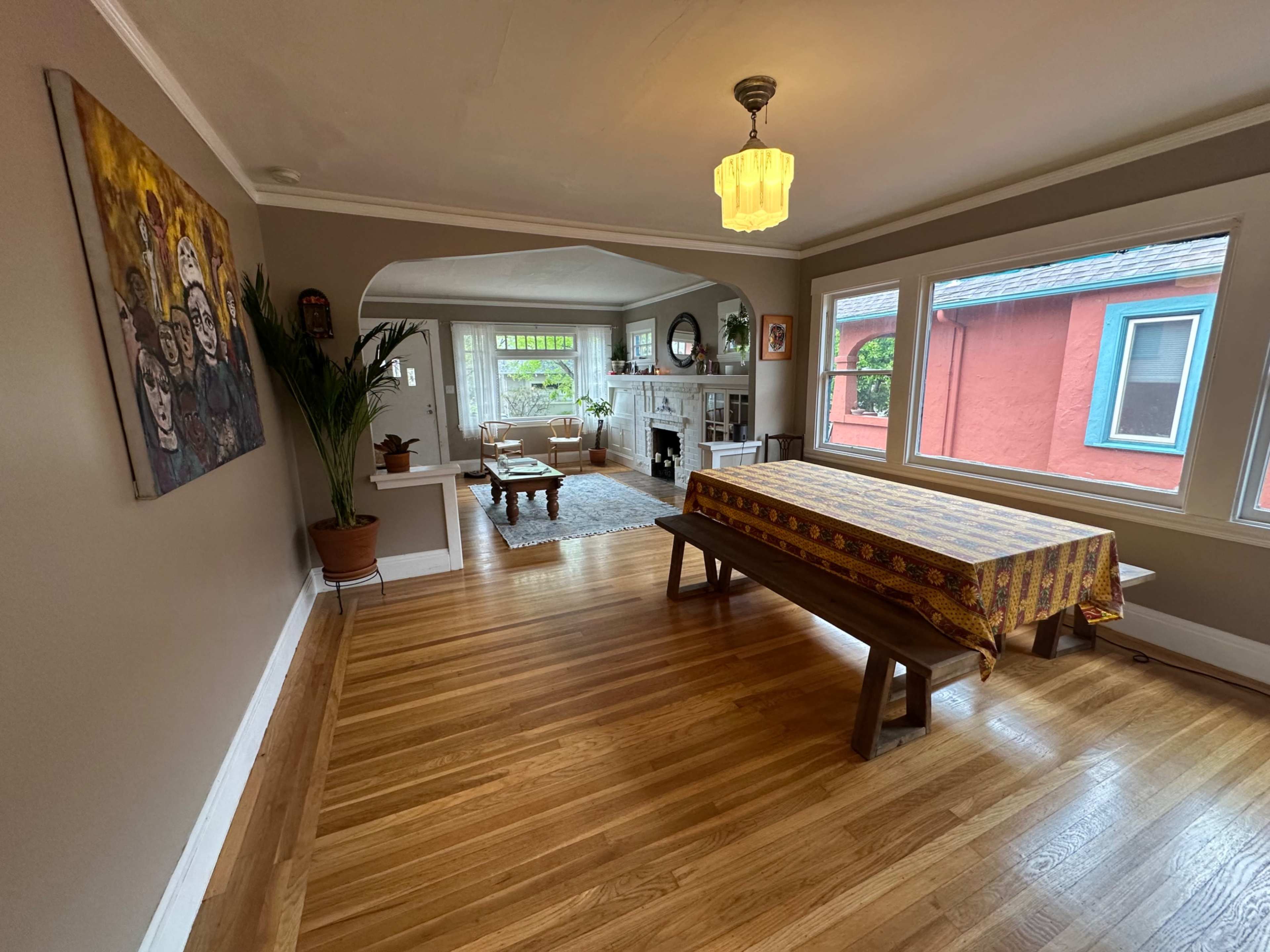 The image shows a dining area with a long wooden table covered with a patterned tablecloth, hardwood floors, and large windows overlooking a living space adorned with a plant and decorative items.