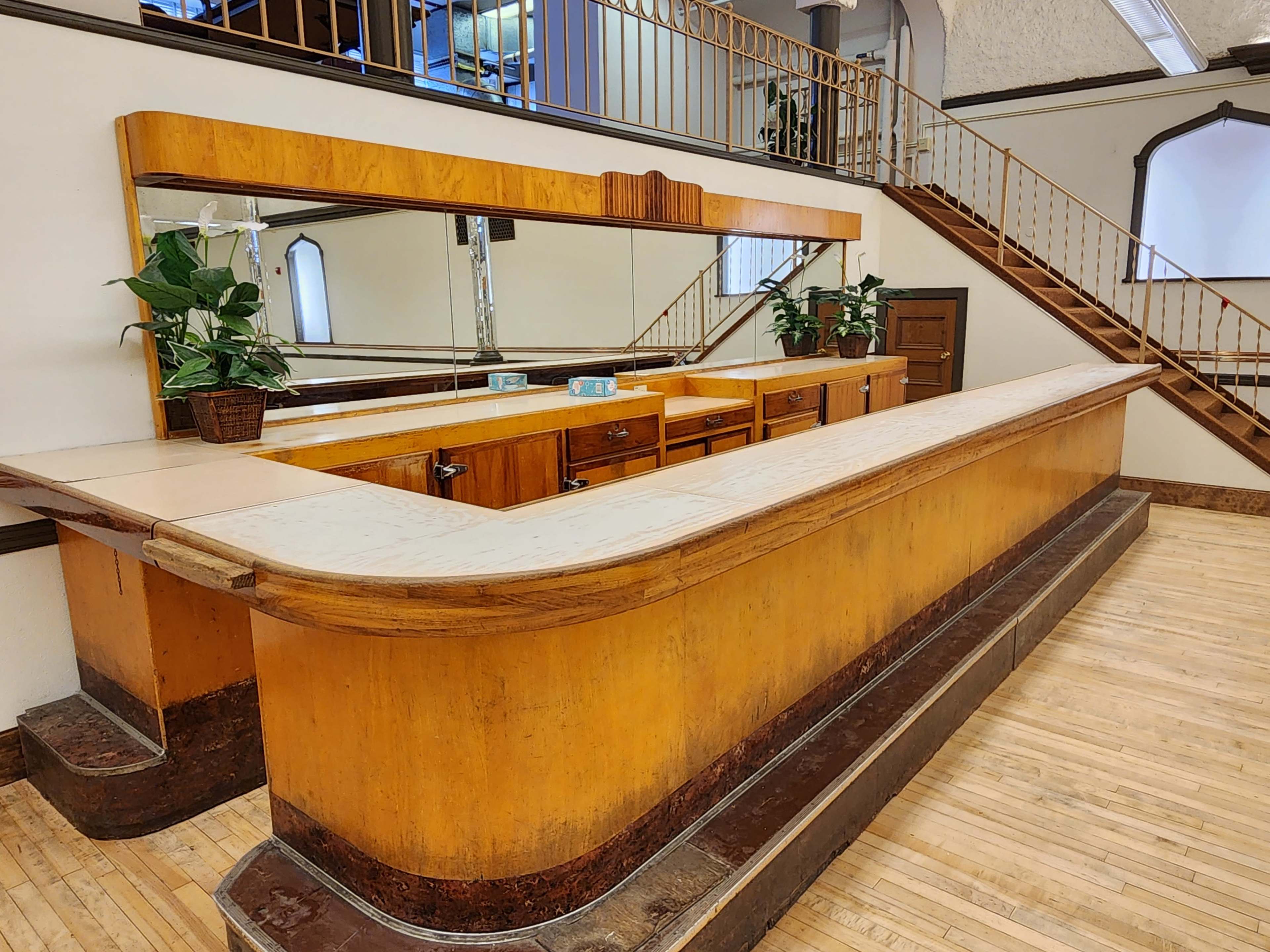 A wooden reception counter with a mirrored back and potted plants, situated in a space with a wooden floor and a staircase in the background.