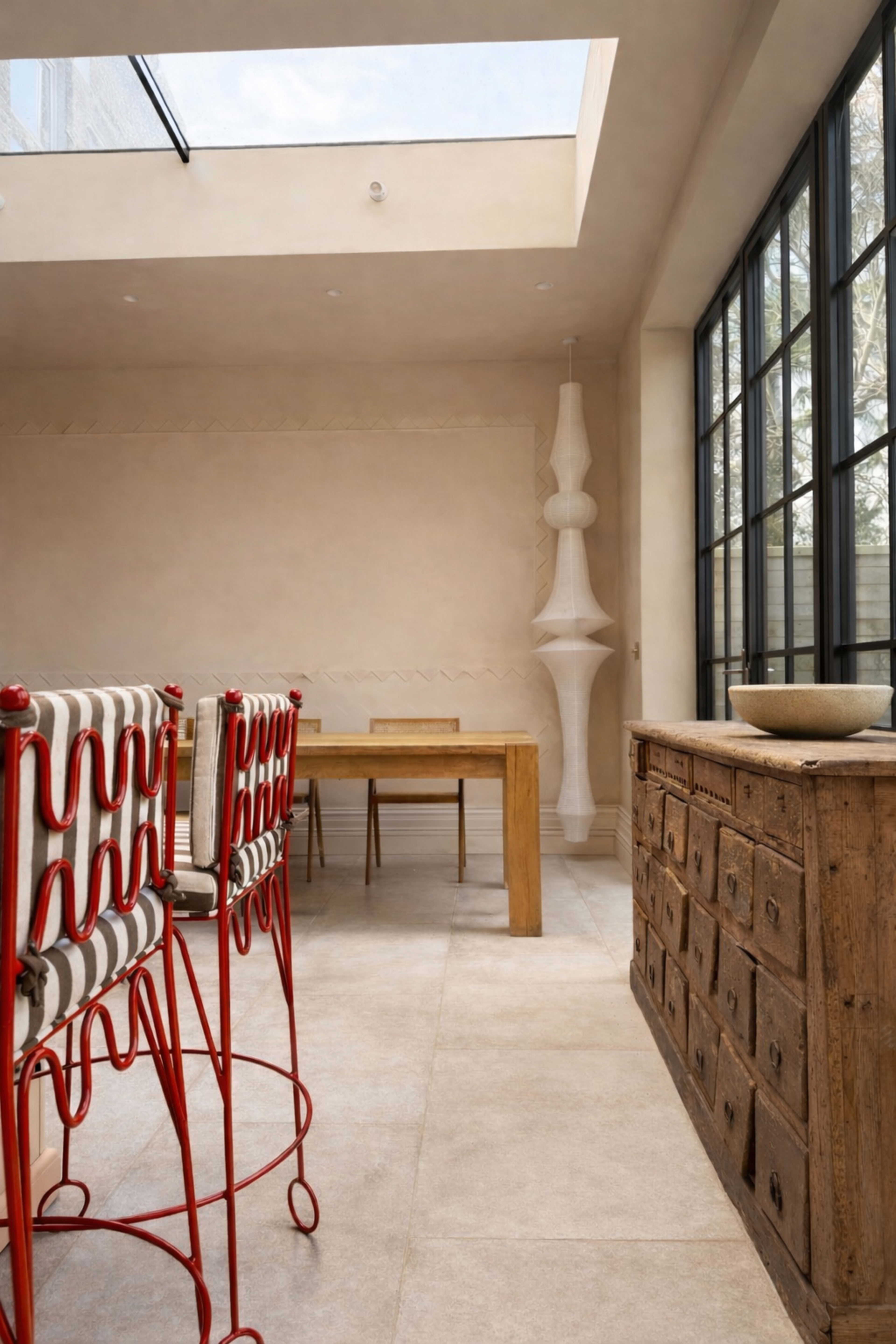 The image shows a modern dining area featuring wooden furniture, red bar stools, and large windows that allow natural light to enter the space.