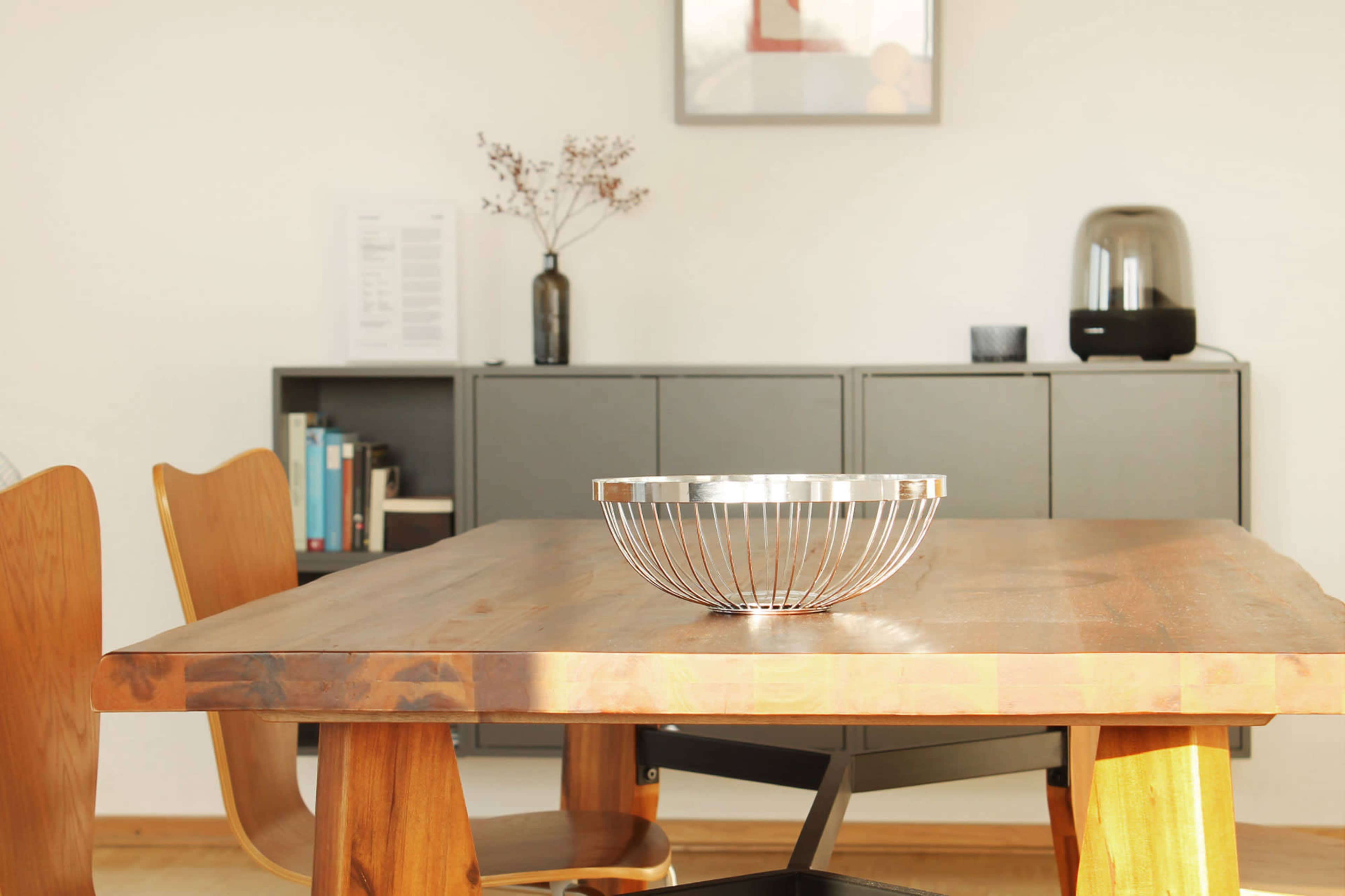 A wooden dining table with a metal fruit bowl is positioned in a modern interior, surrounded by two wooden chairs and a cabinet in the background.