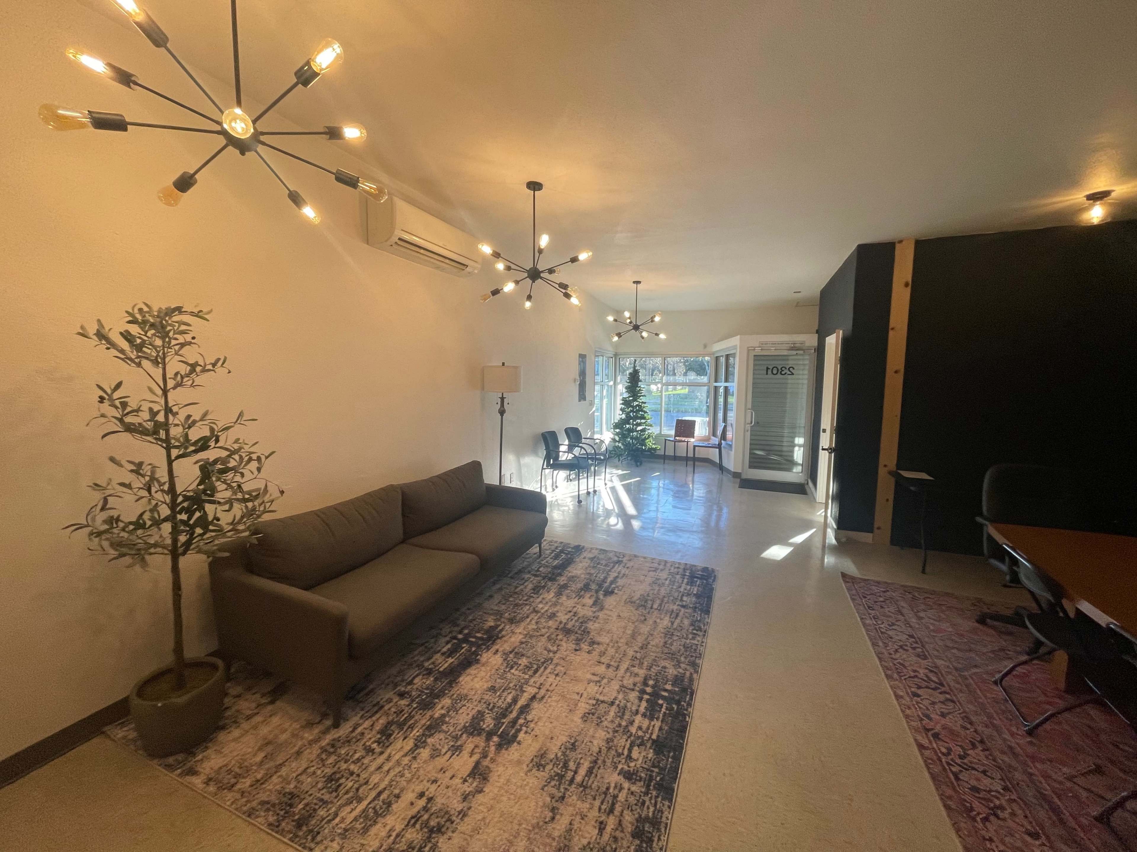 The scene features a spacious room with a sofa, plants, and modern light fixtures, leading to a glass door that opens to an outdoor view.