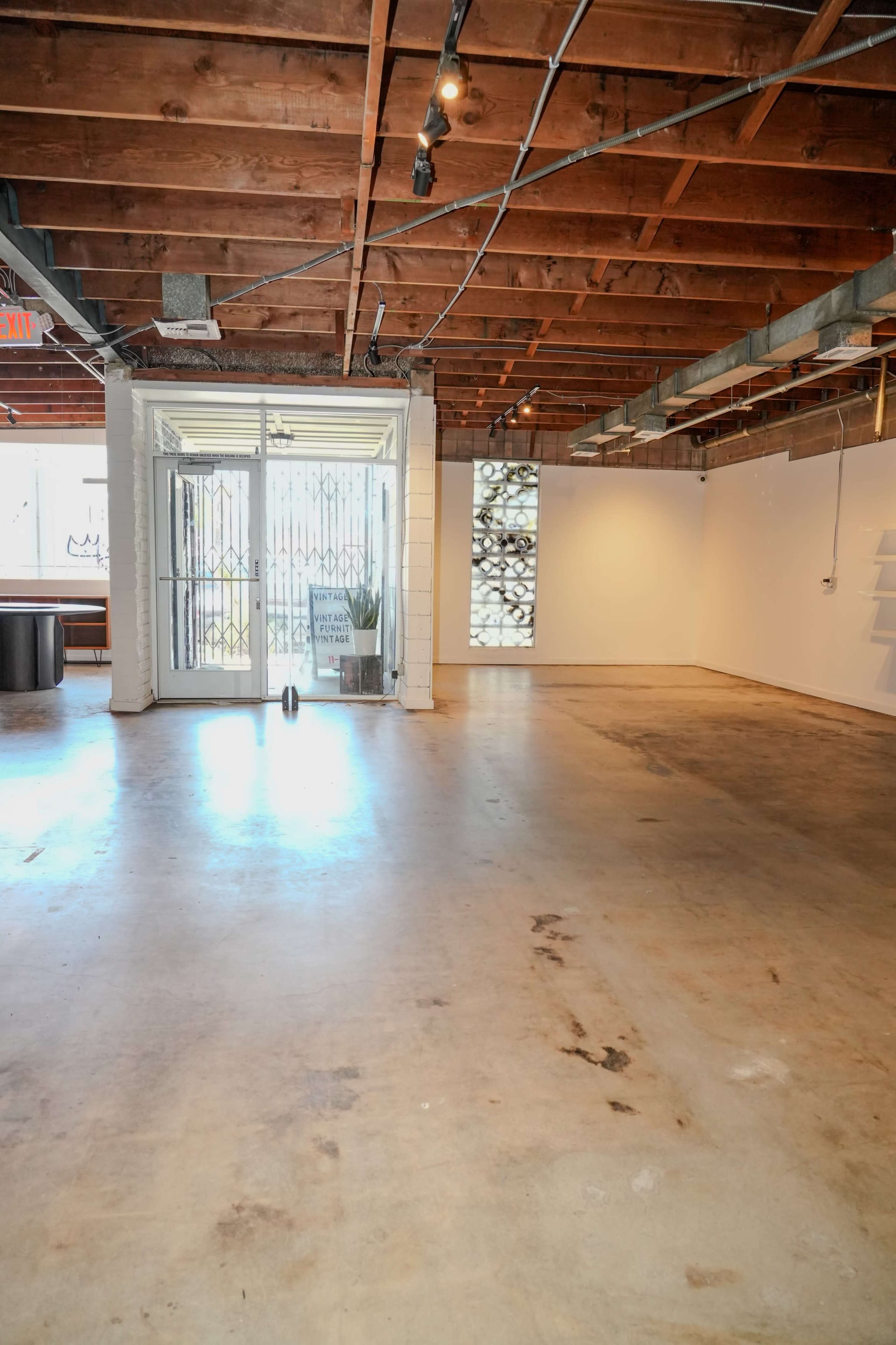 A spacious, empty room with exposed wooden beams, concrete flooring, and large windows that open to the outside.