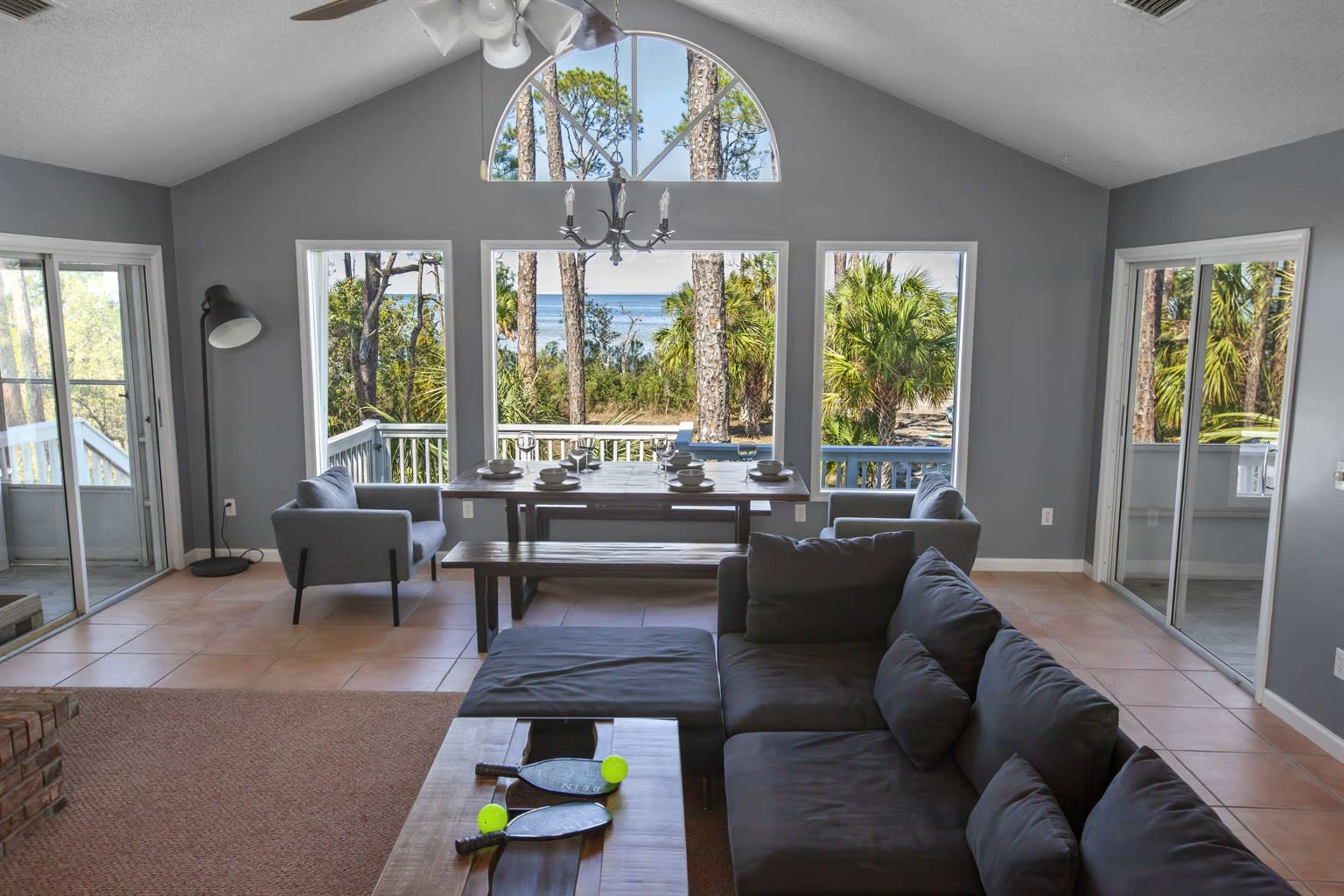 The image shows a spacious, well-lit living area with a large window offering a view of trees and a body of water.