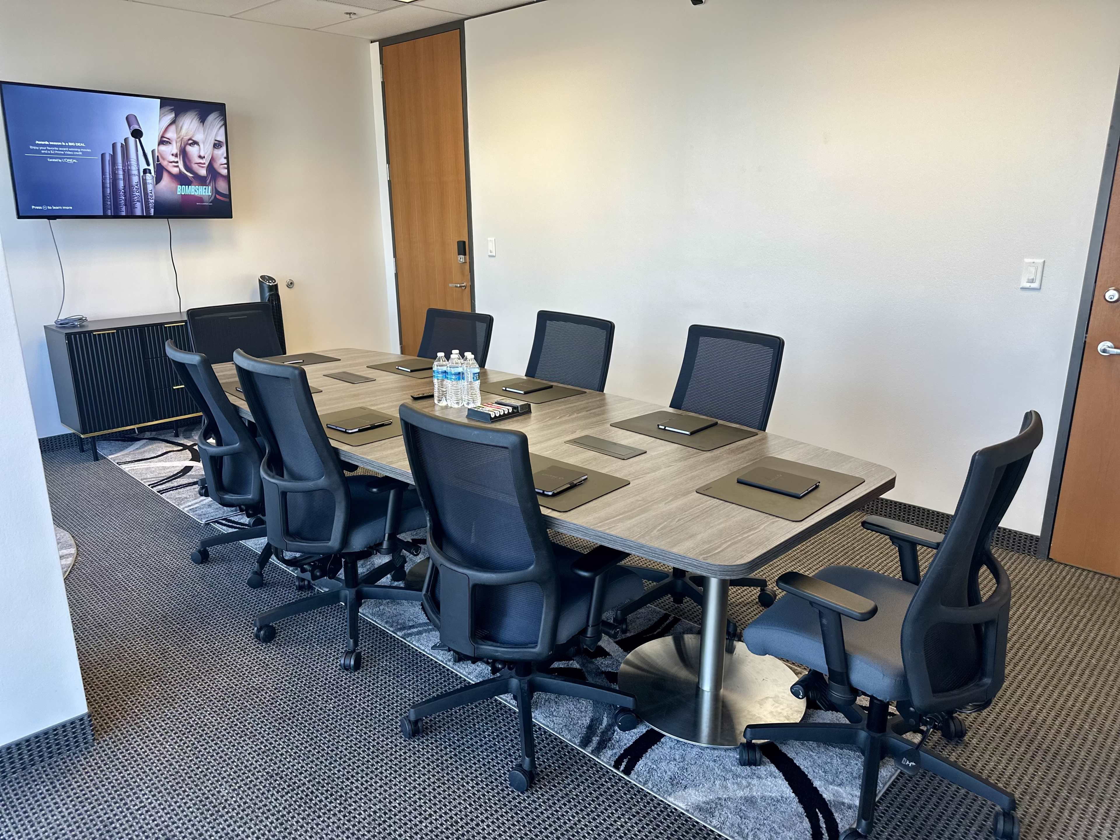 A conference room features a long table surrounded by black chairs, with a wall-mounted screen displaying a promotional image.