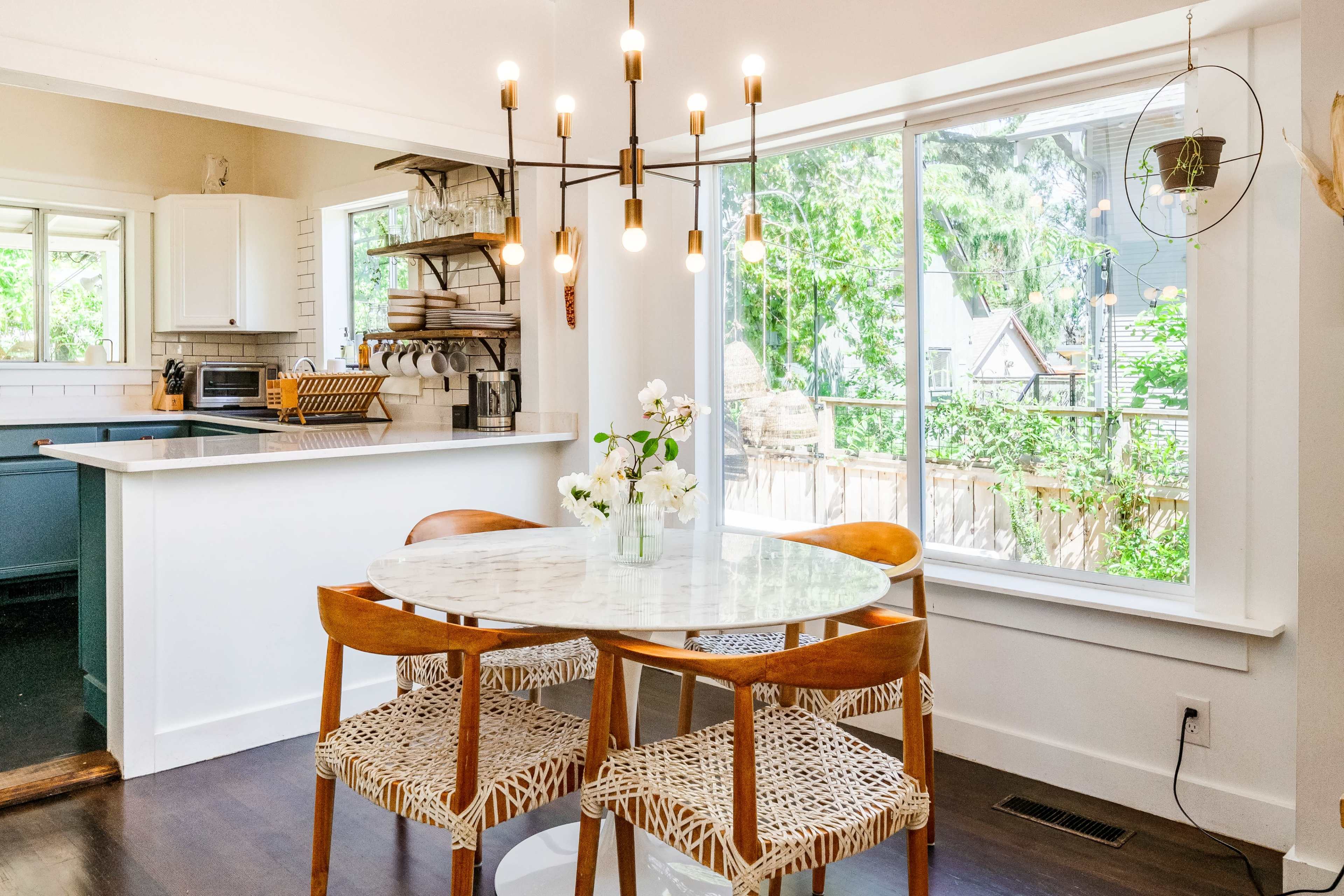 A dining area features a round marble table surrounded by three woven chairs, illuminated by a modern chandelier, with a view of a garden through large windows.