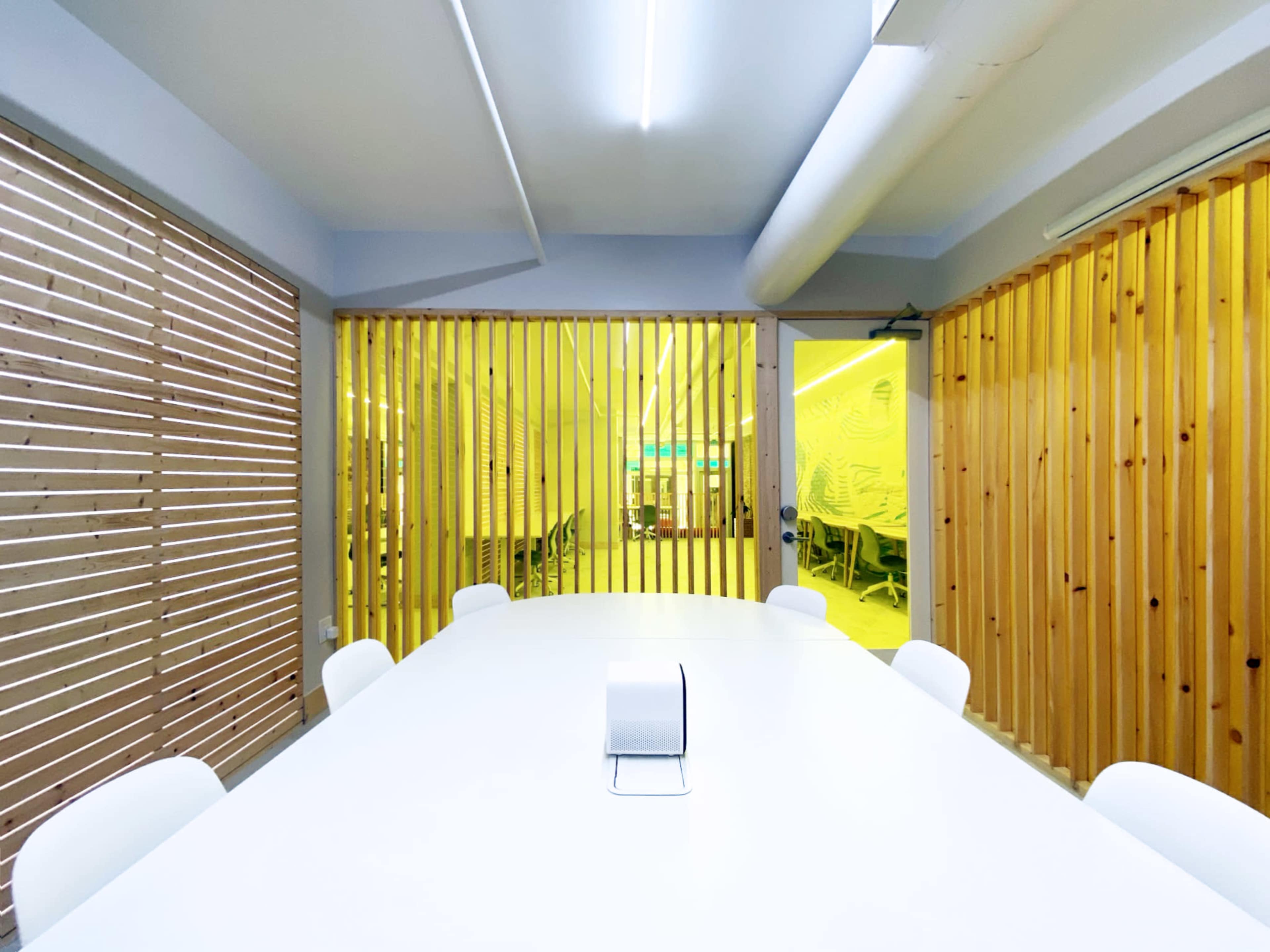 The image shows a modern meeting room with a long white table surrounded by chairs and warm wood paneling.