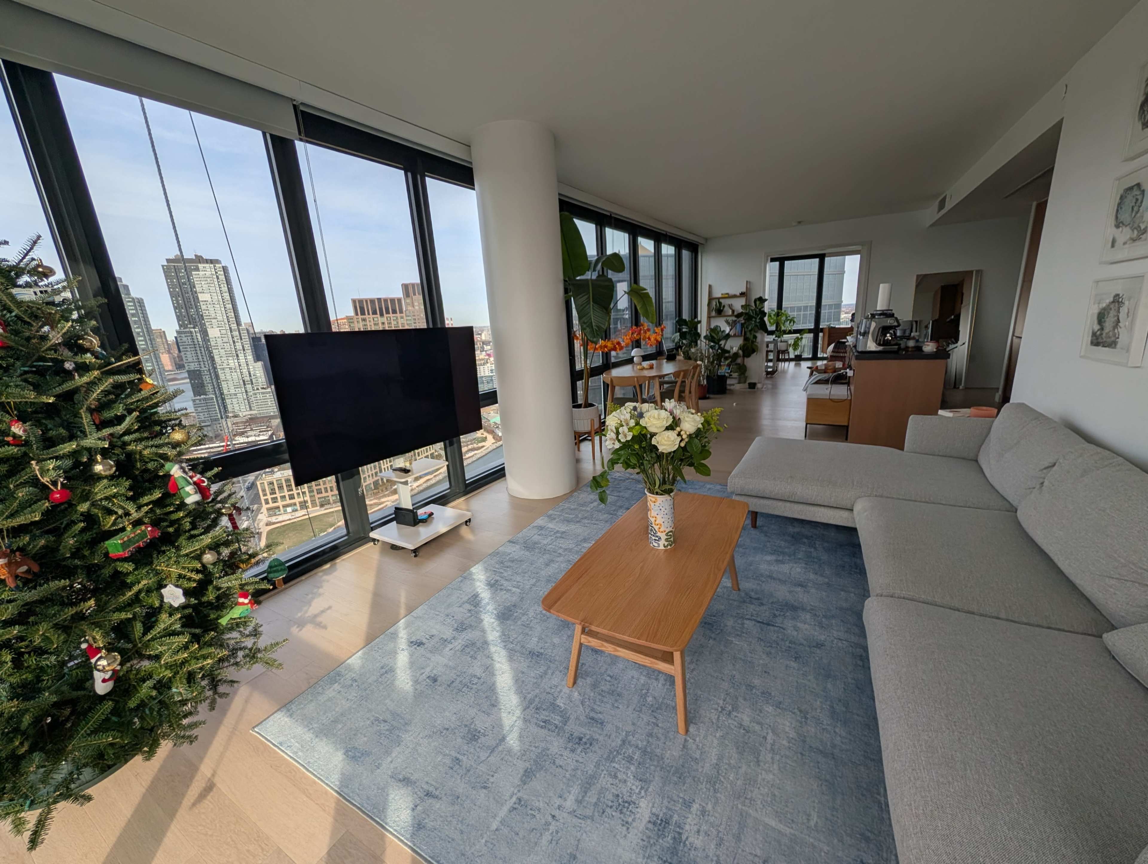 The image shows a modern living room with a large window, a light gray sofa, a wooden coffee table, a flat-screen TV, and a Christmas tree in a corner.