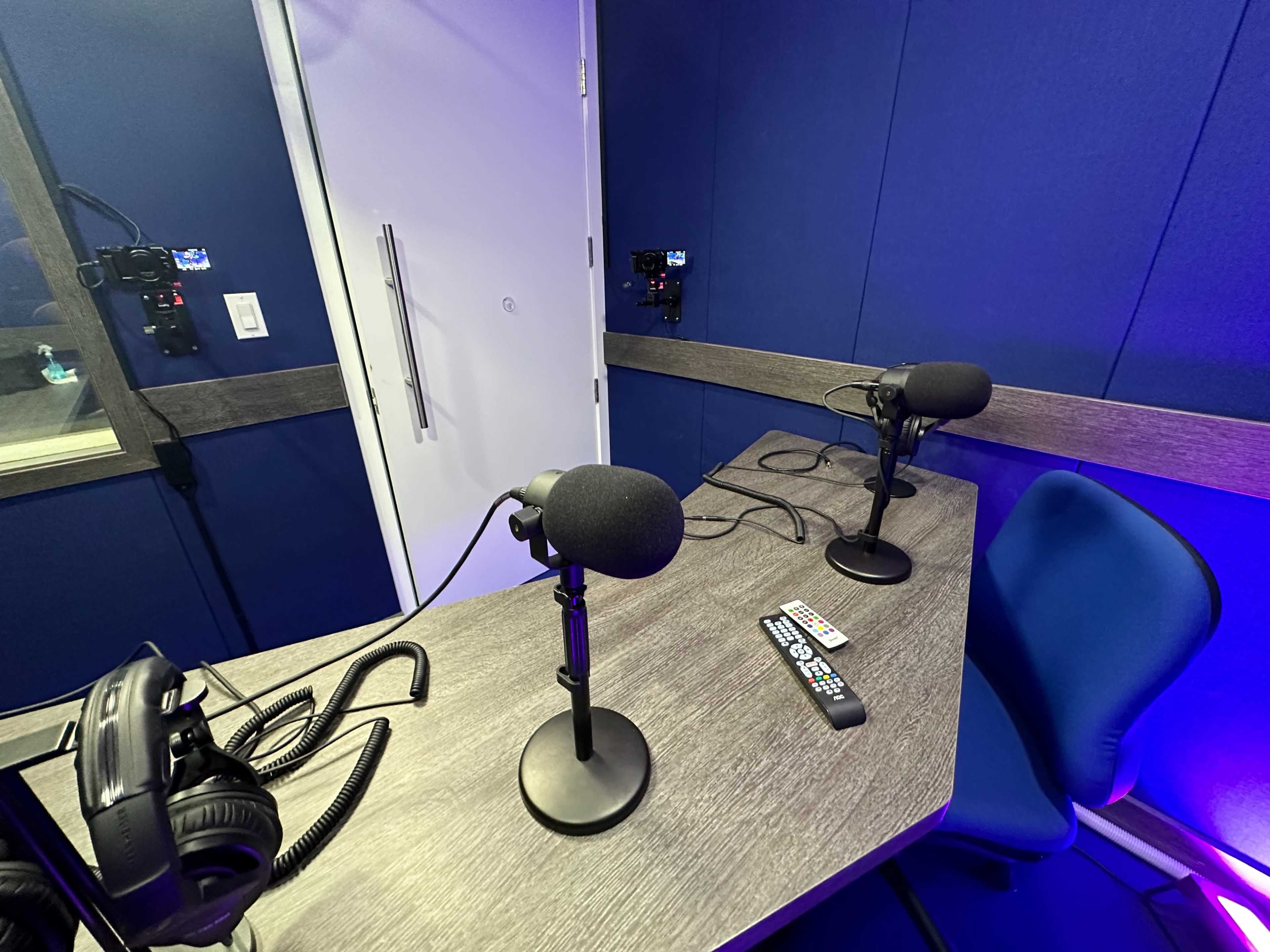 A recording studio is shown with two microphones on a table, headphones, and remote controls nearby, set against a blue wall.