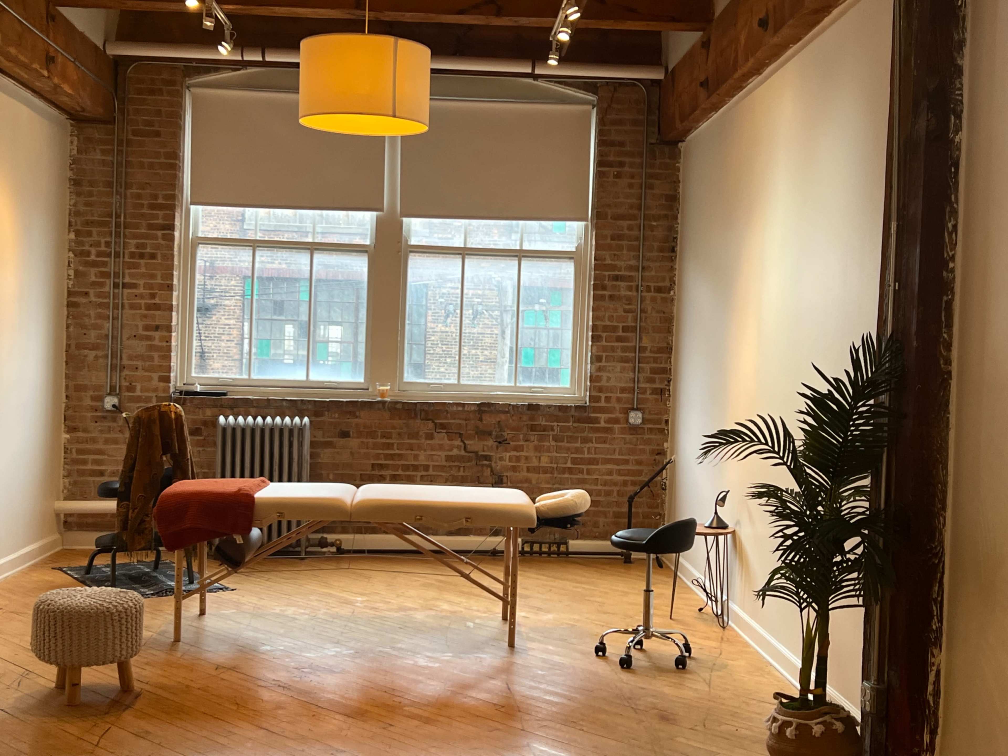 The image shows a well-lit therapy room with a massage table, a chair, plants, and large windows overlooking a brick wall.