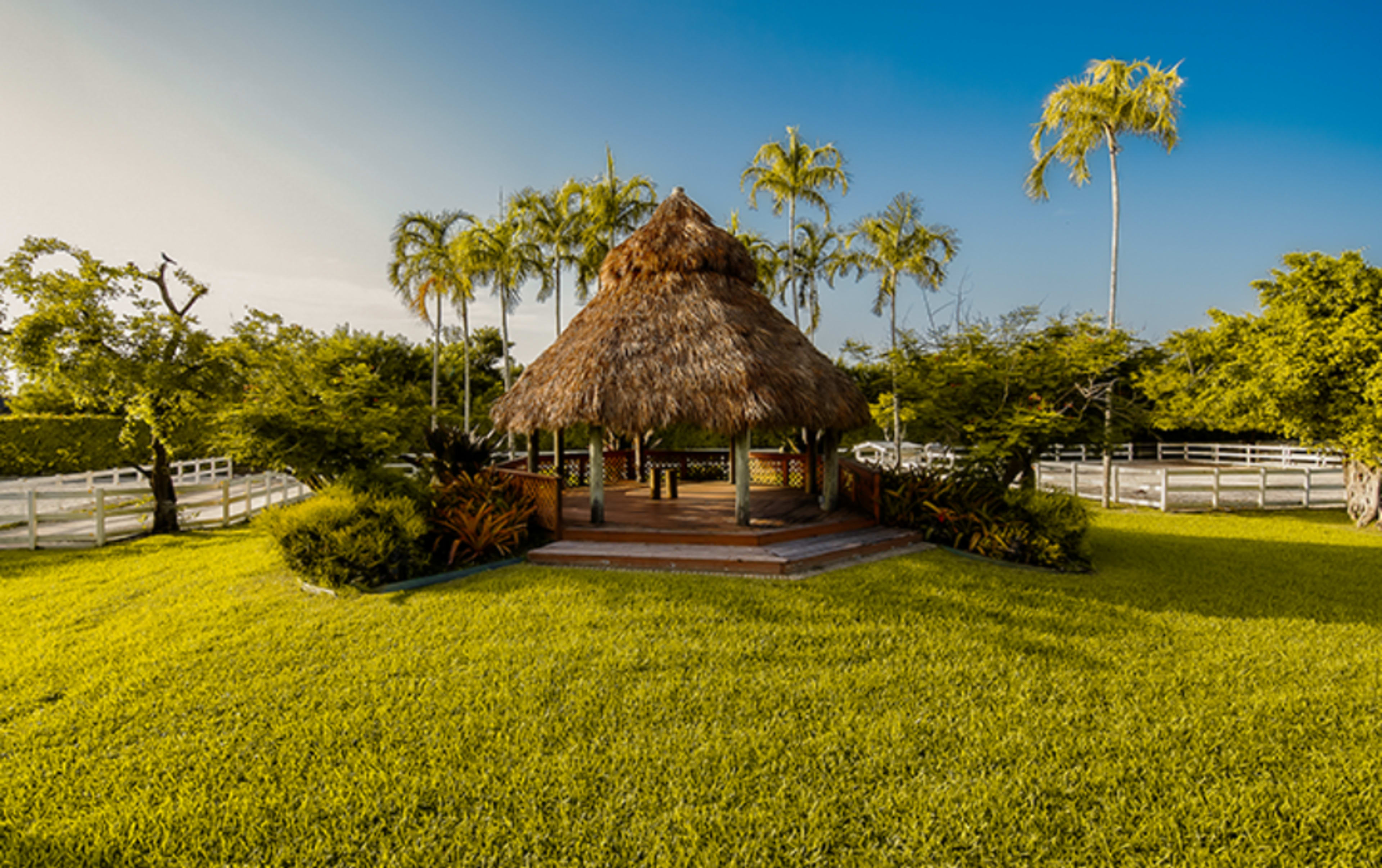 A thatched gazebo is positioned on a manicured lawn surrounded by tropical trees and a white fence under a clear blue sky.