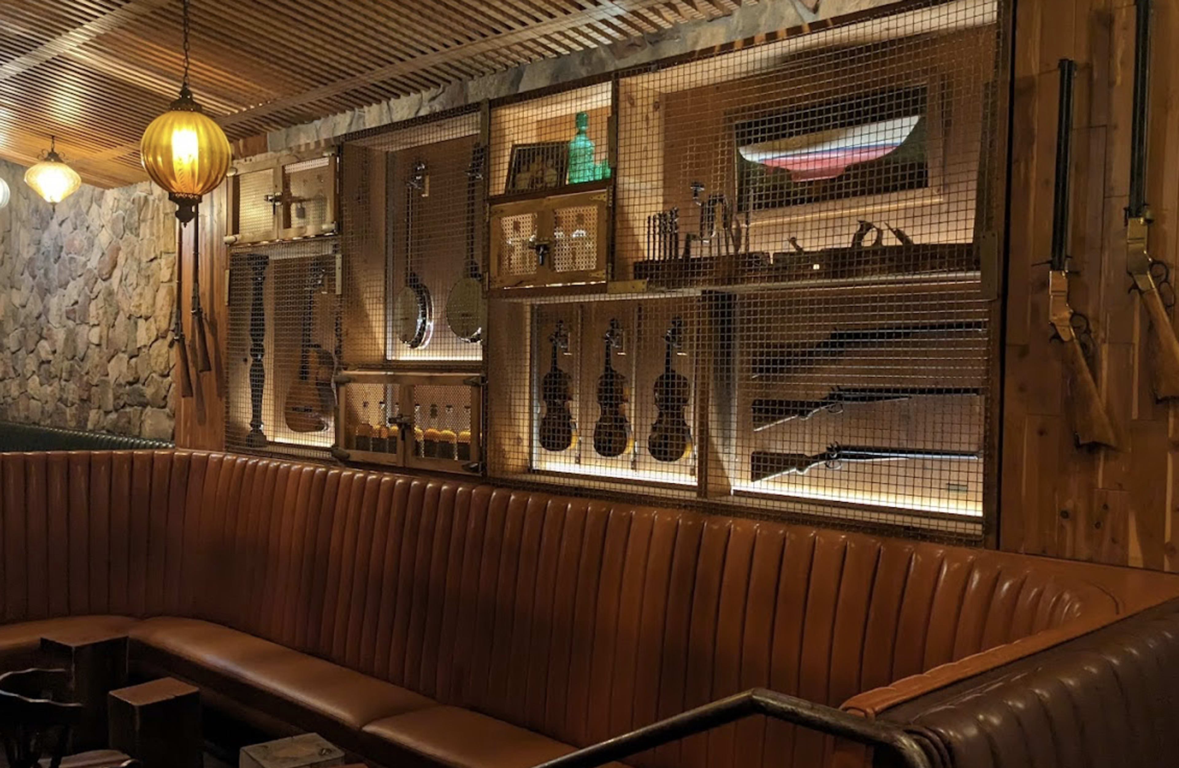 The image features a cozy restaurant interior with a wooden wall displaying various musical instruments and vintage firearms behind a wire mesh, alongside a leather banquette seating area.