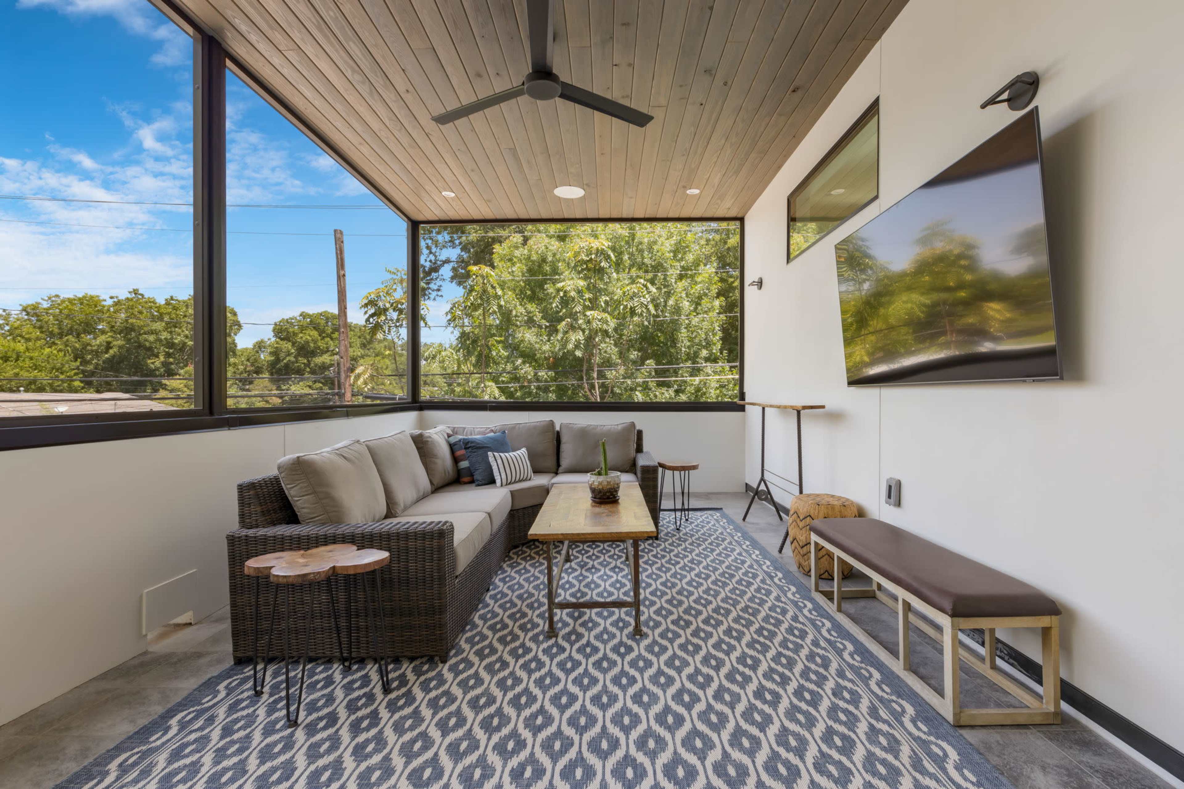 The image depicts a modern, enclosed patio featuring a comfortable seating area with a sofa, coffee table, and a wall-mounted television, surrounded by large windows that provide ample natural light.