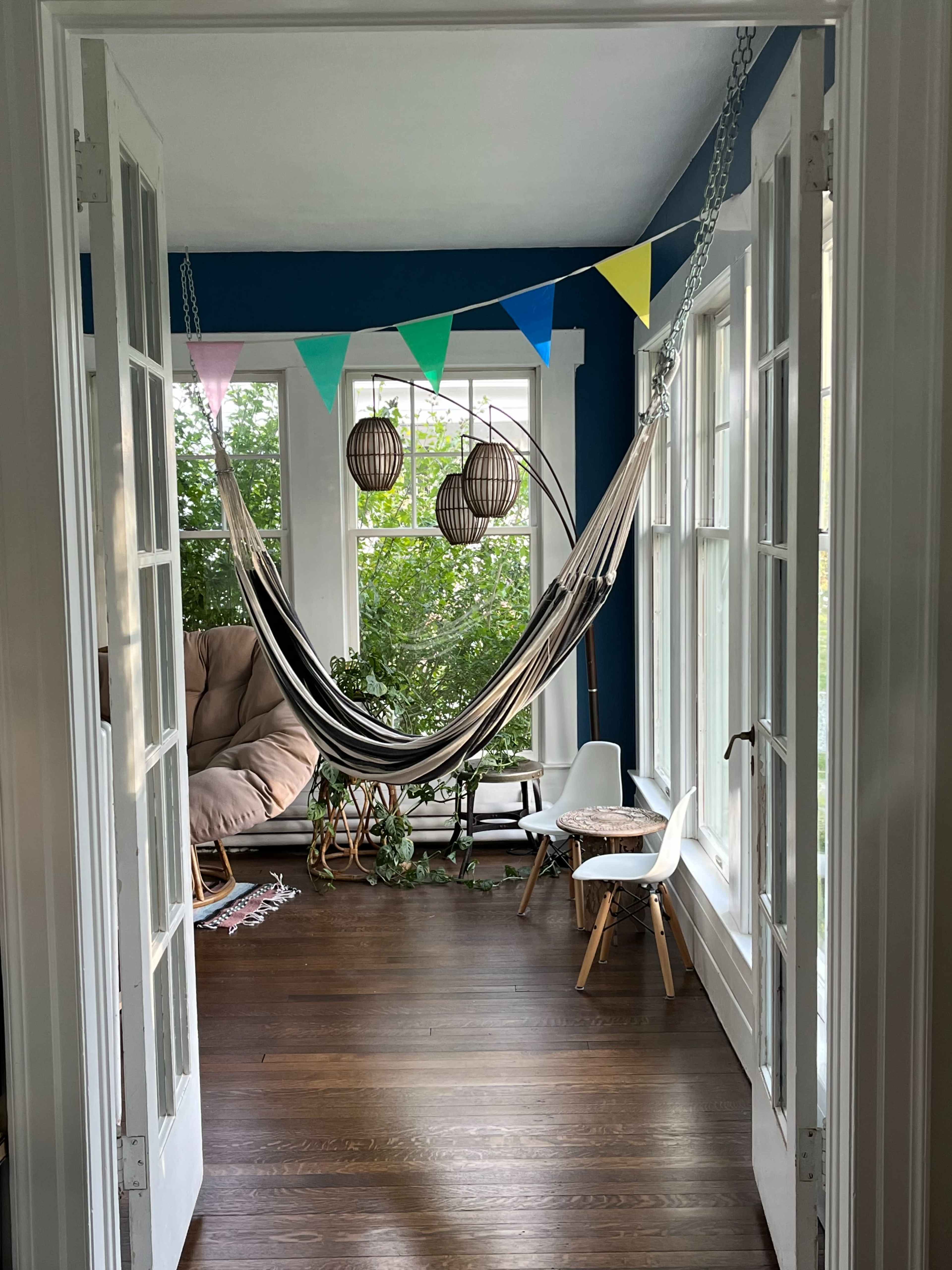 A hammock is suspended between two walls in a bright room filled with plants and decorated with colorful bunting and hanging light fixtures.