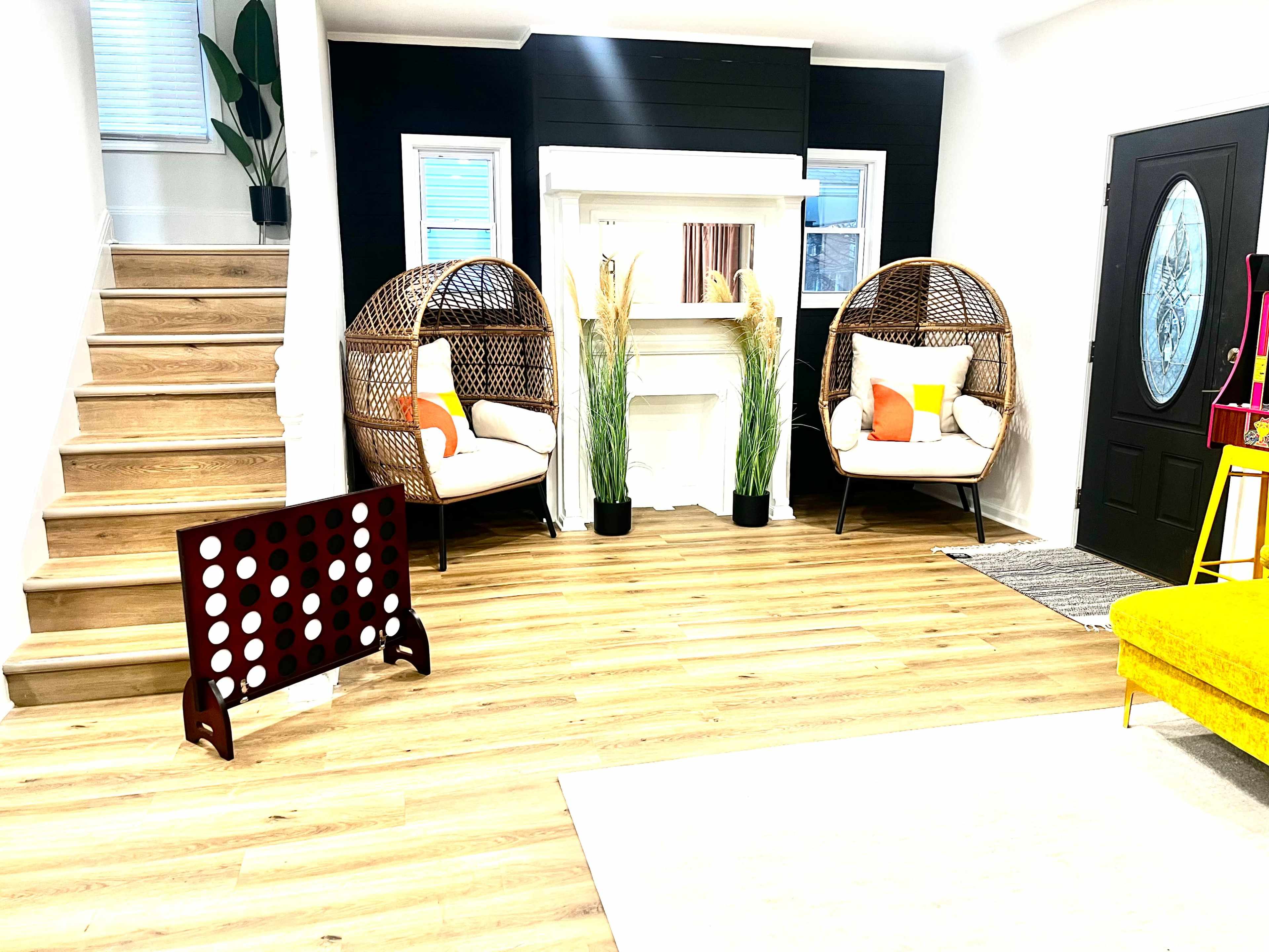 The room features a staircase, two round wicker chairs with cushions, a decorative fireplace, tall plants, and a large Connect Four game in the foreground.