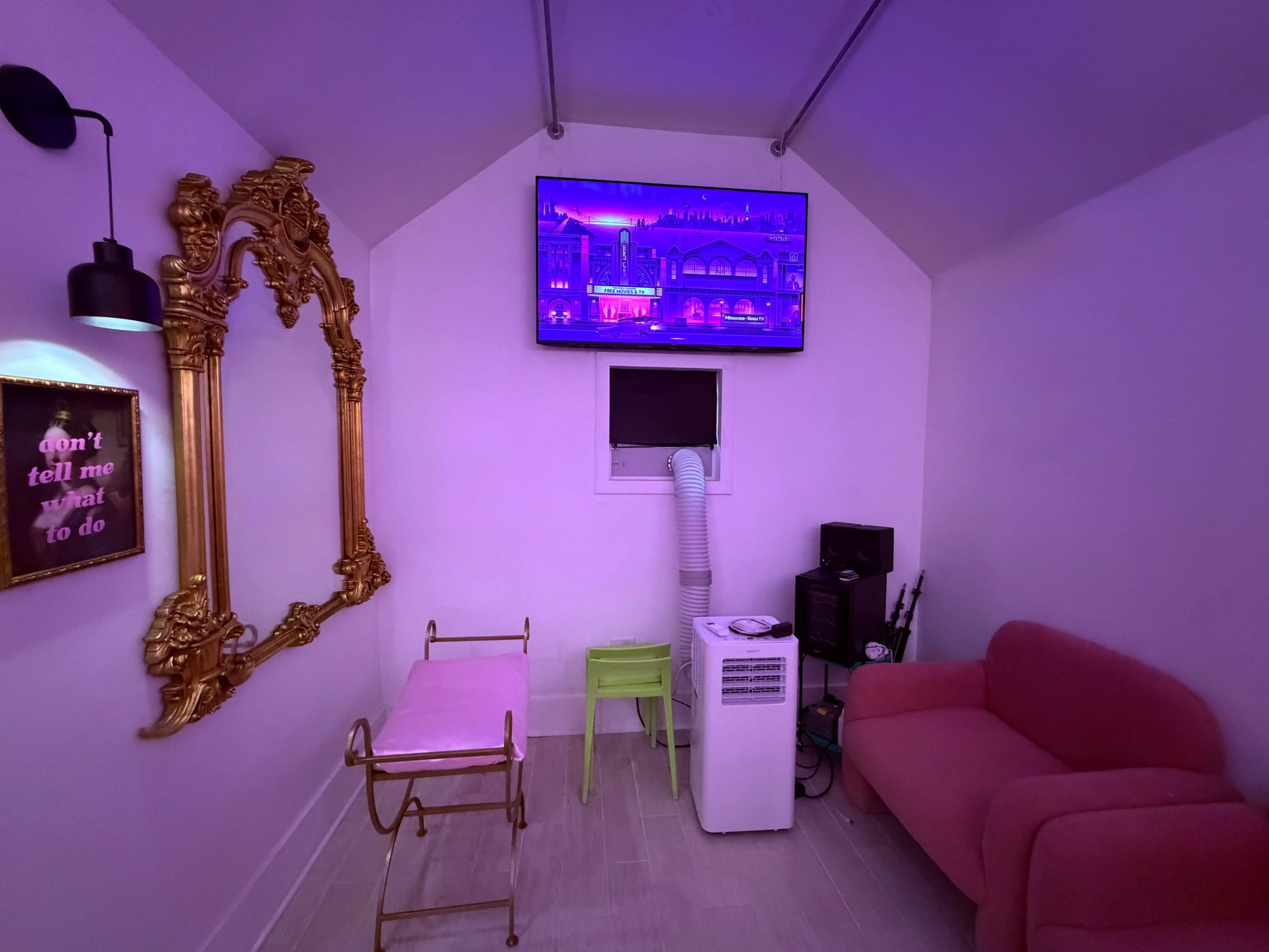 The image shows a small, brightly colored room with a pink couch, a green chair, a white air conditioning unit, and a large mirror on the wall, all illuminated by purple light.