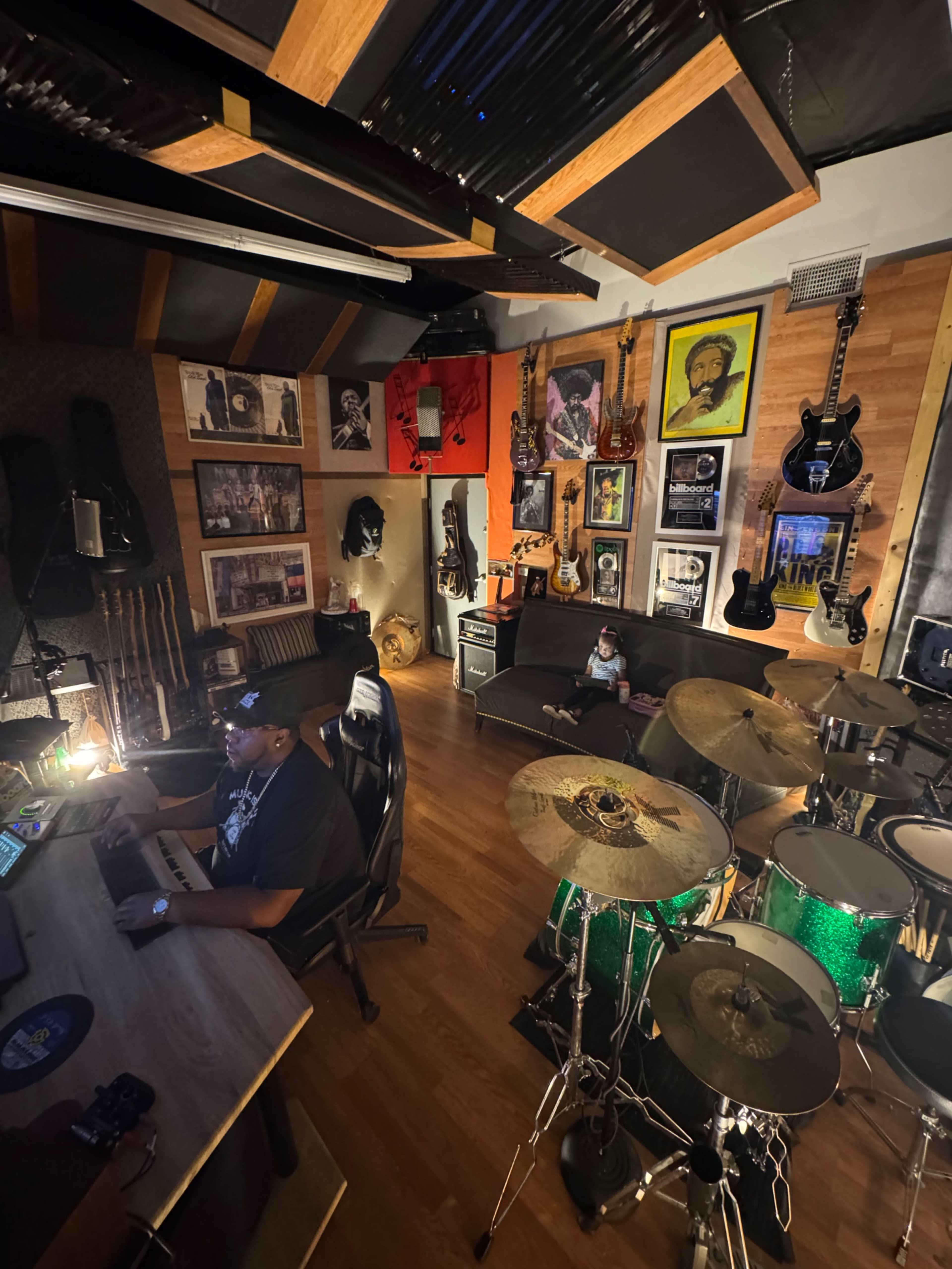 A musician works at a computer in a home studio surrounded by various instruments, artwork, and musical memorabilia.