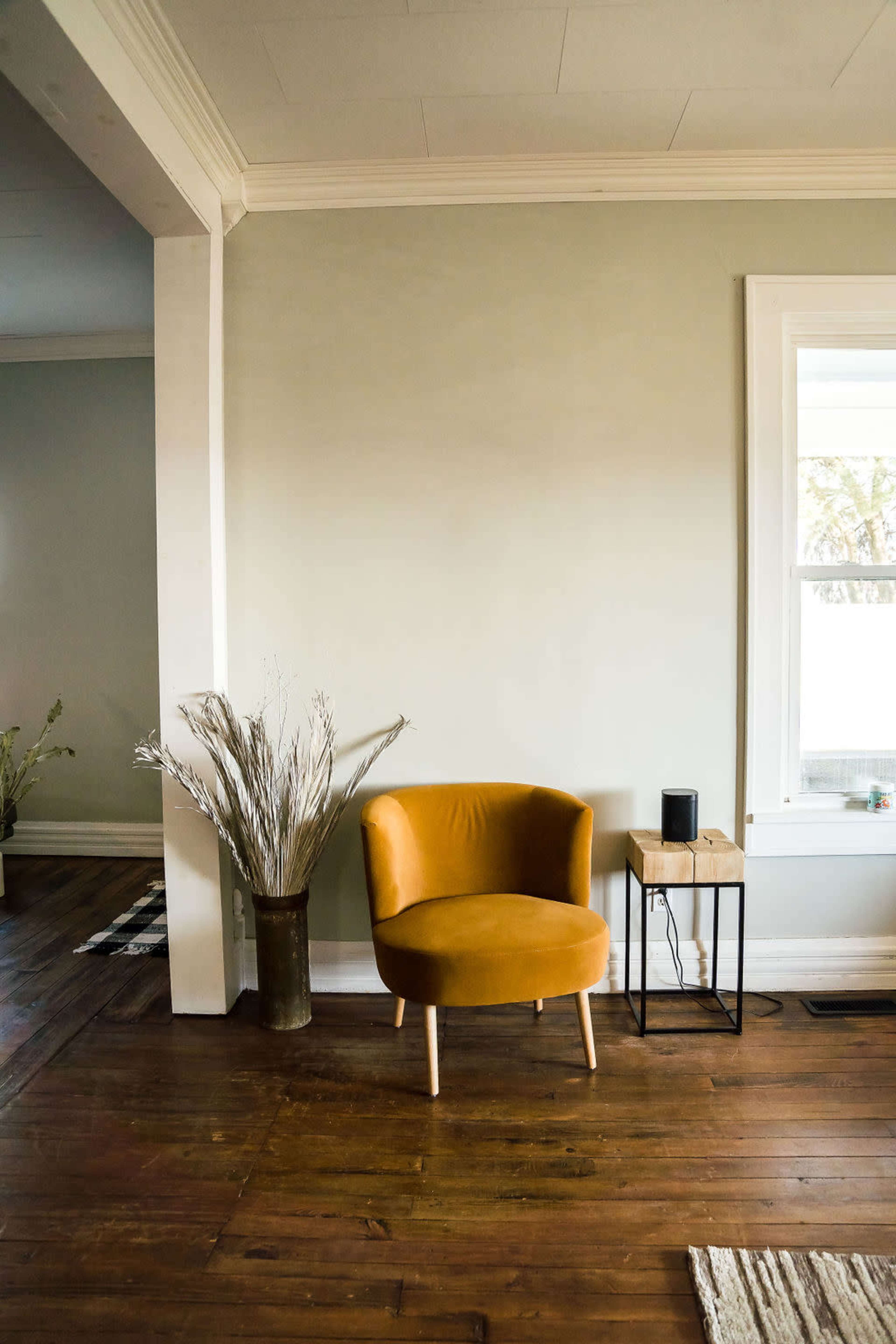 A rounded mustard-colored chair is positioned next to a small wooden side table in a room with light green walls and wooden flooring.