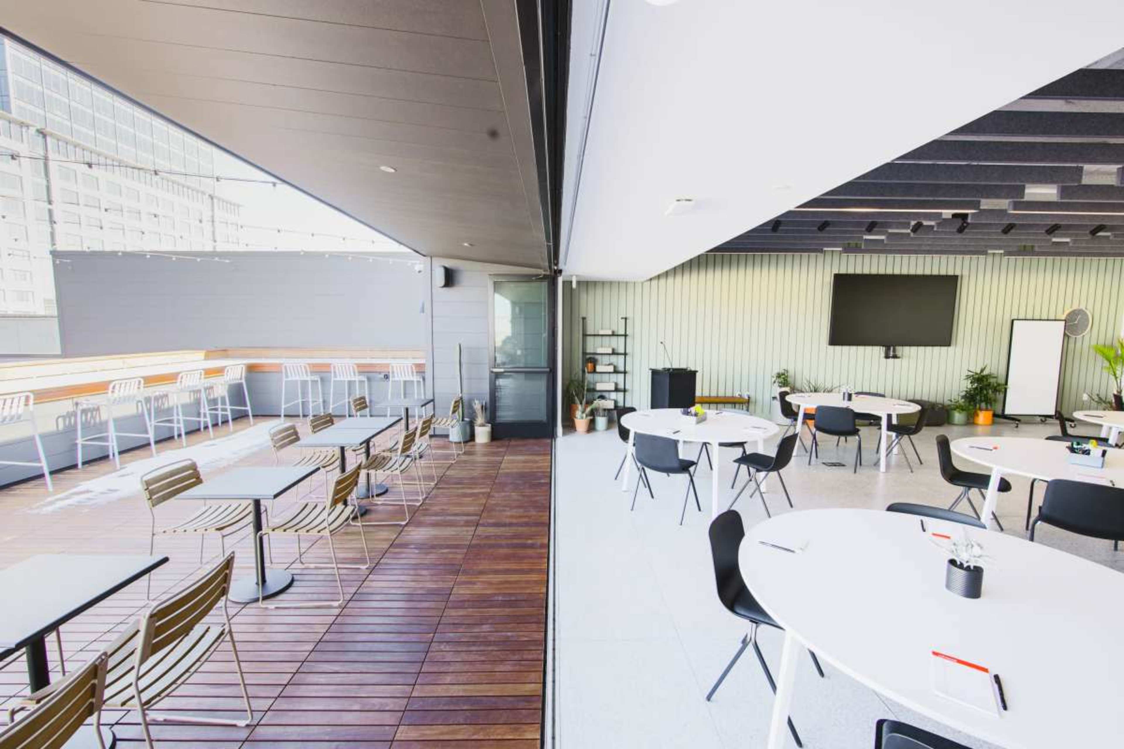 The image shows a conference space divided into two sections: one side features an outdoor wooden deck with seating, while the other side has a carpeted indoor area with circular tables and a large screen.
