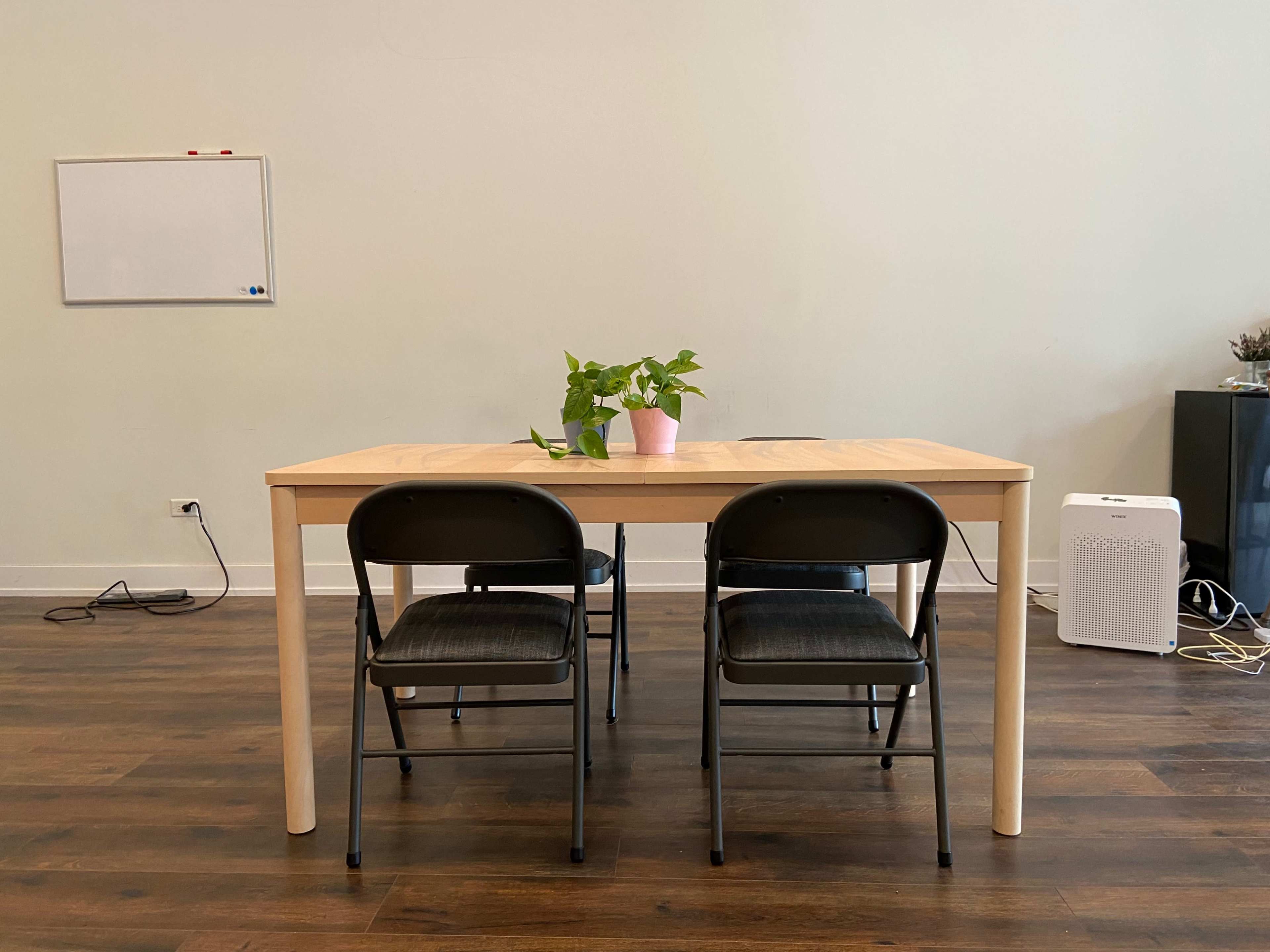 A wooden table is set in a minimalist room with two black chairs, a small plant in a pink pot, and a whiteboard on the wall.