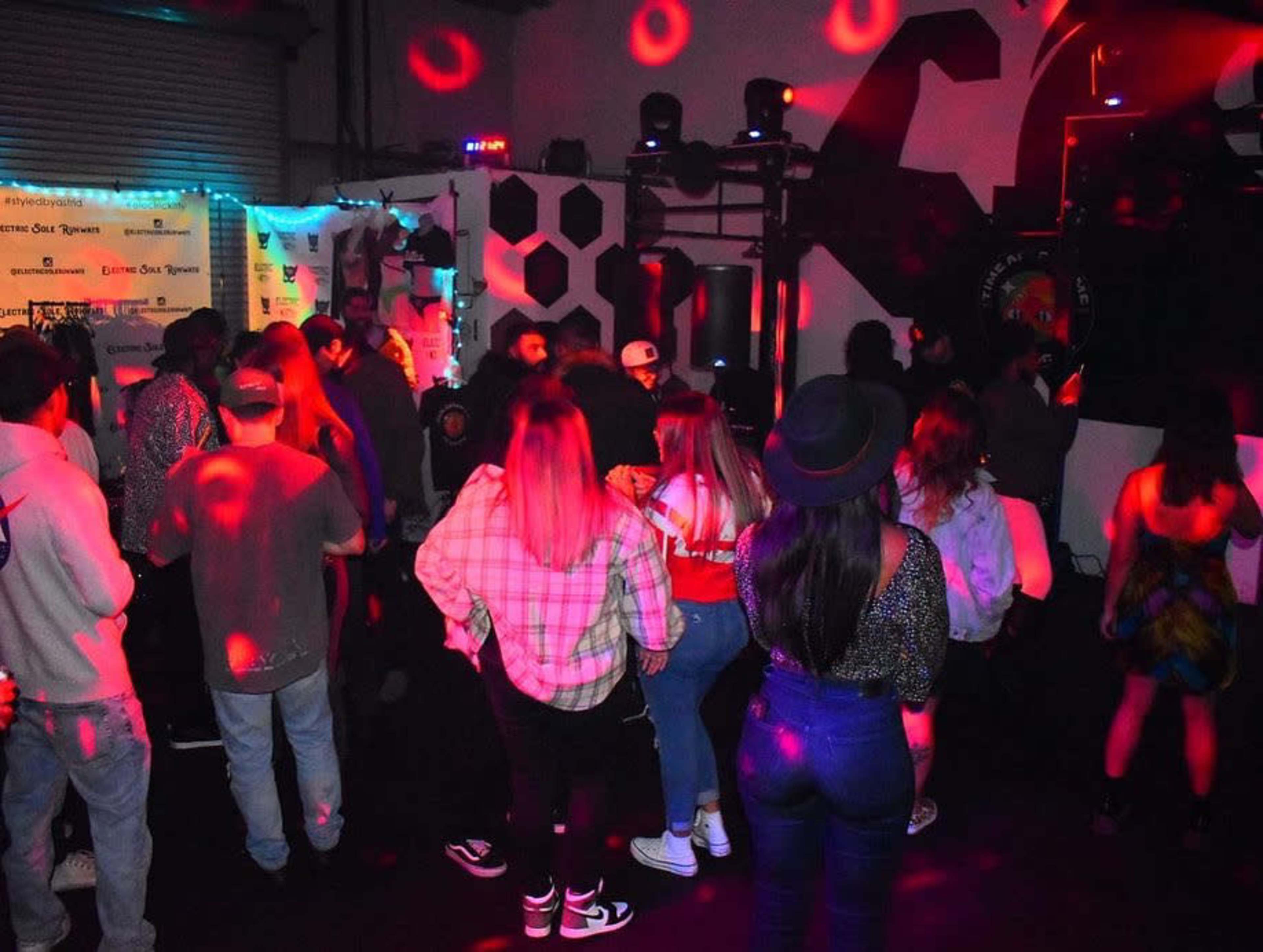 A group of people dance in a dimly lit venue with colorful lights and a DJ setup in the background.