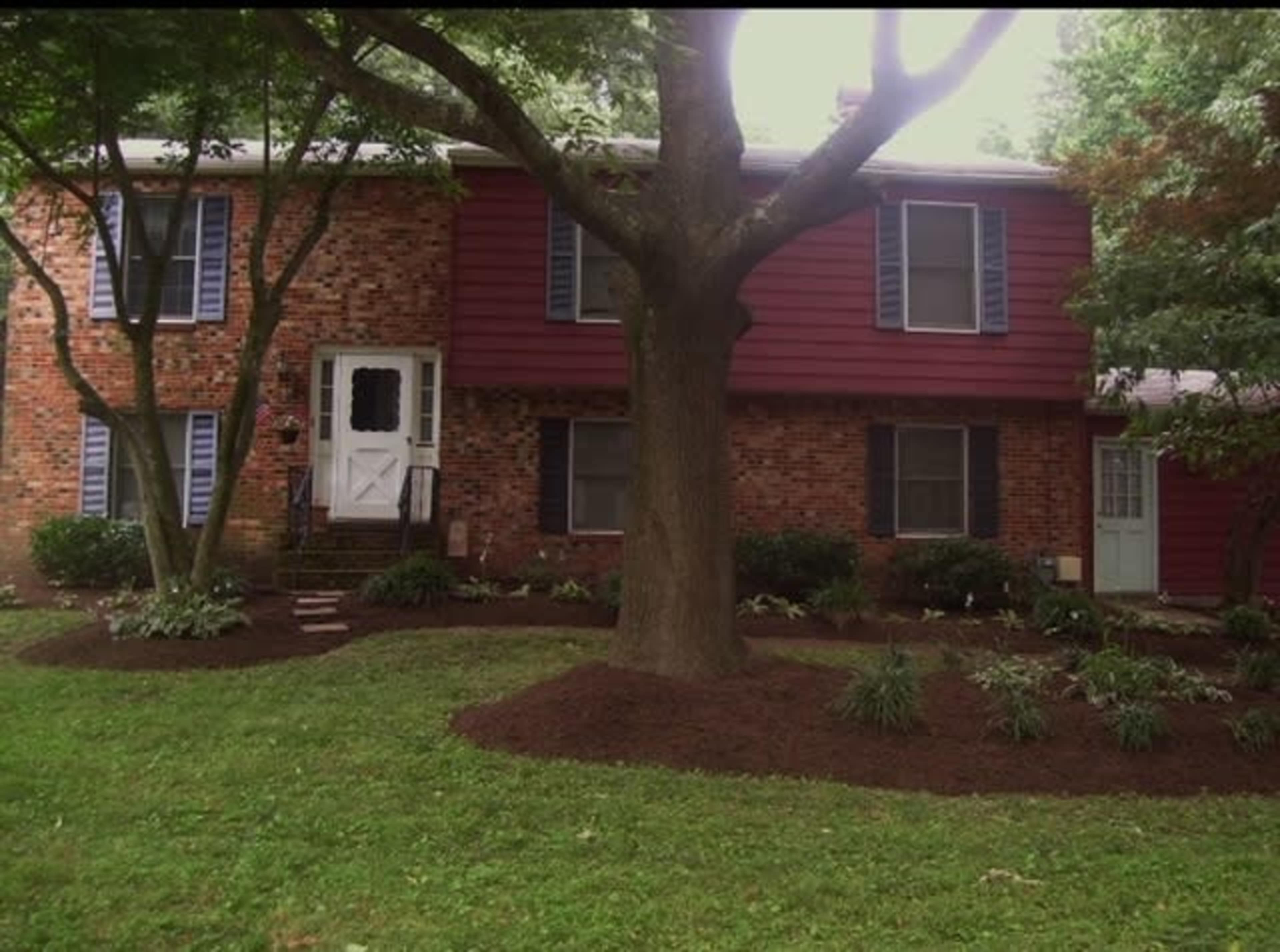 A two-story brick and wood house with blue shutters and a white front door is surrounded by a landscaped yard featuring mulch and small plants.