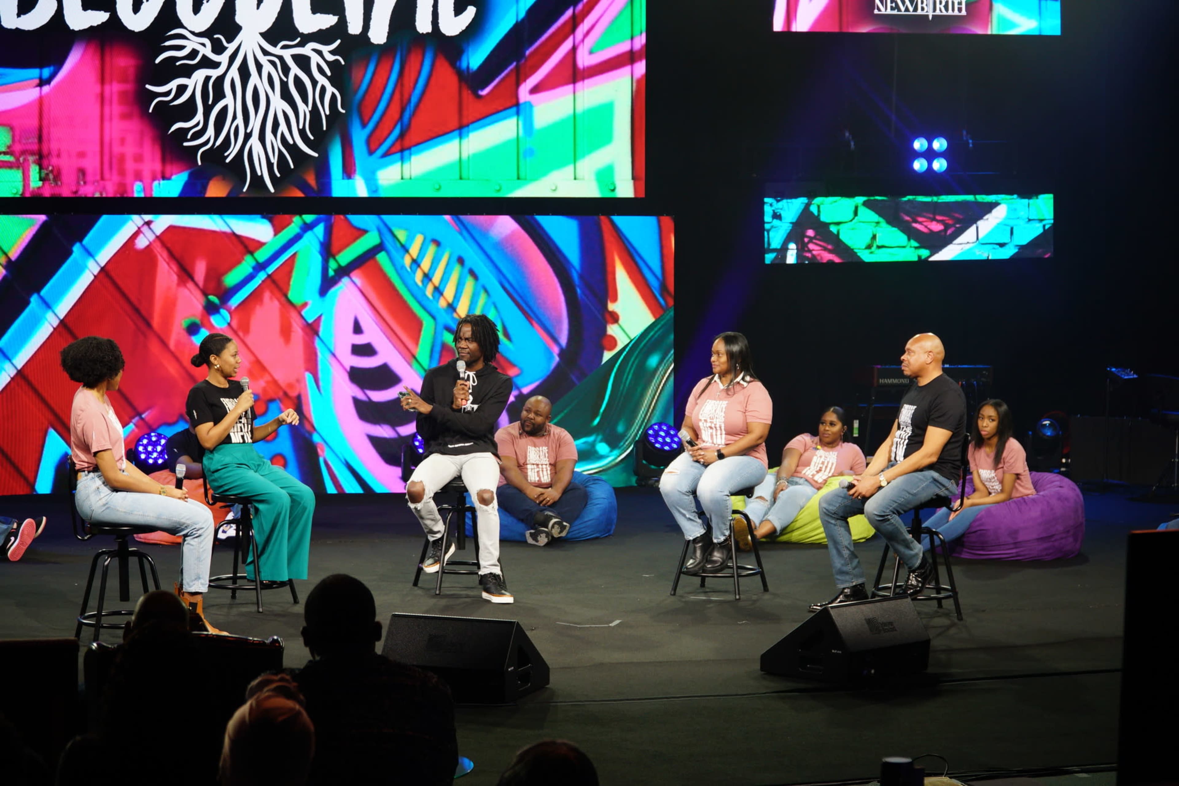 A panel discussion takes place on stage with speakers seated on stools in front of a colorful backdrop, while several participants sit on the floor behind them.