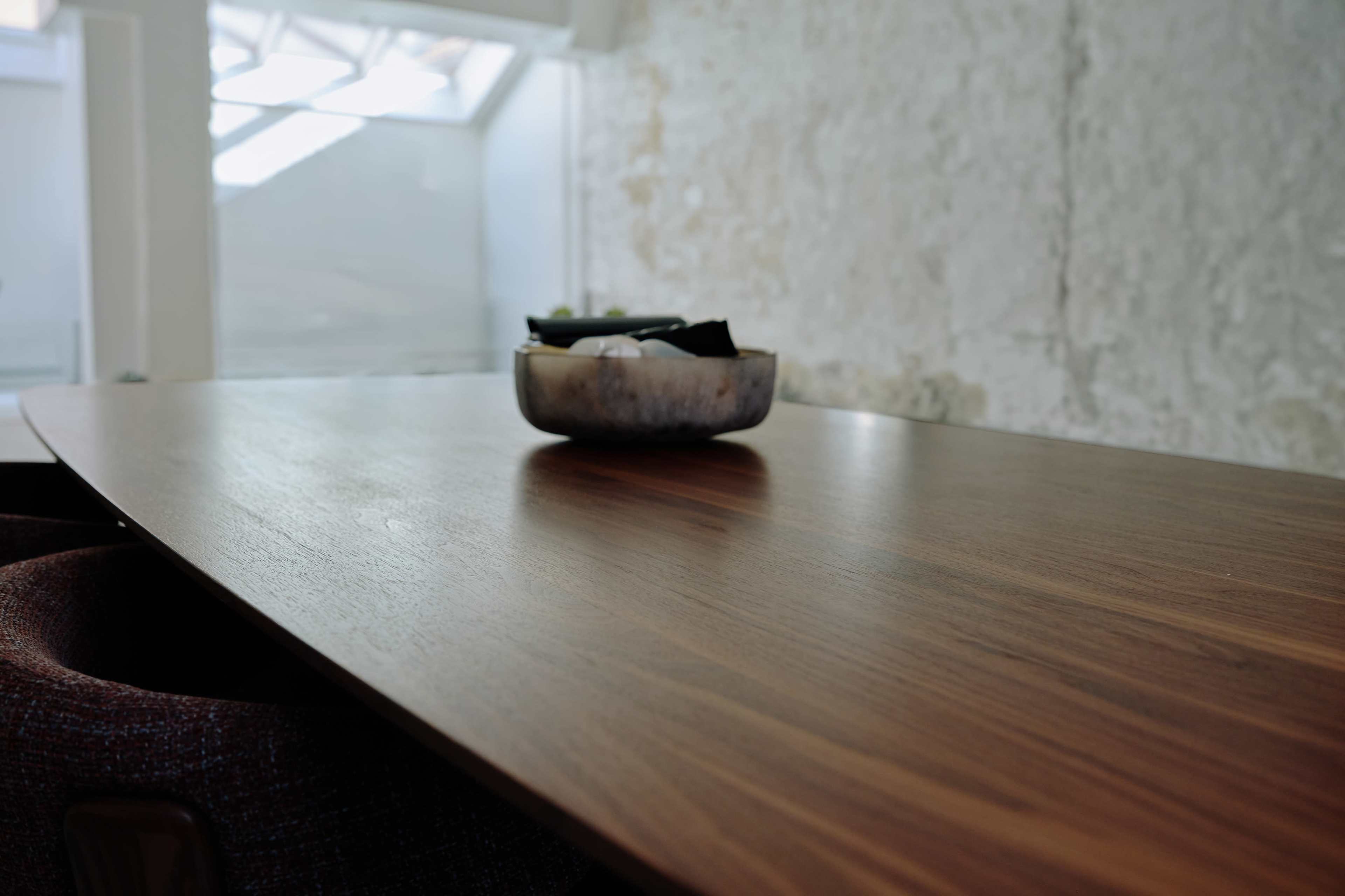 A wooden table with a shallow bowl sitting on top is shown in a well-lit room with a stone wall.