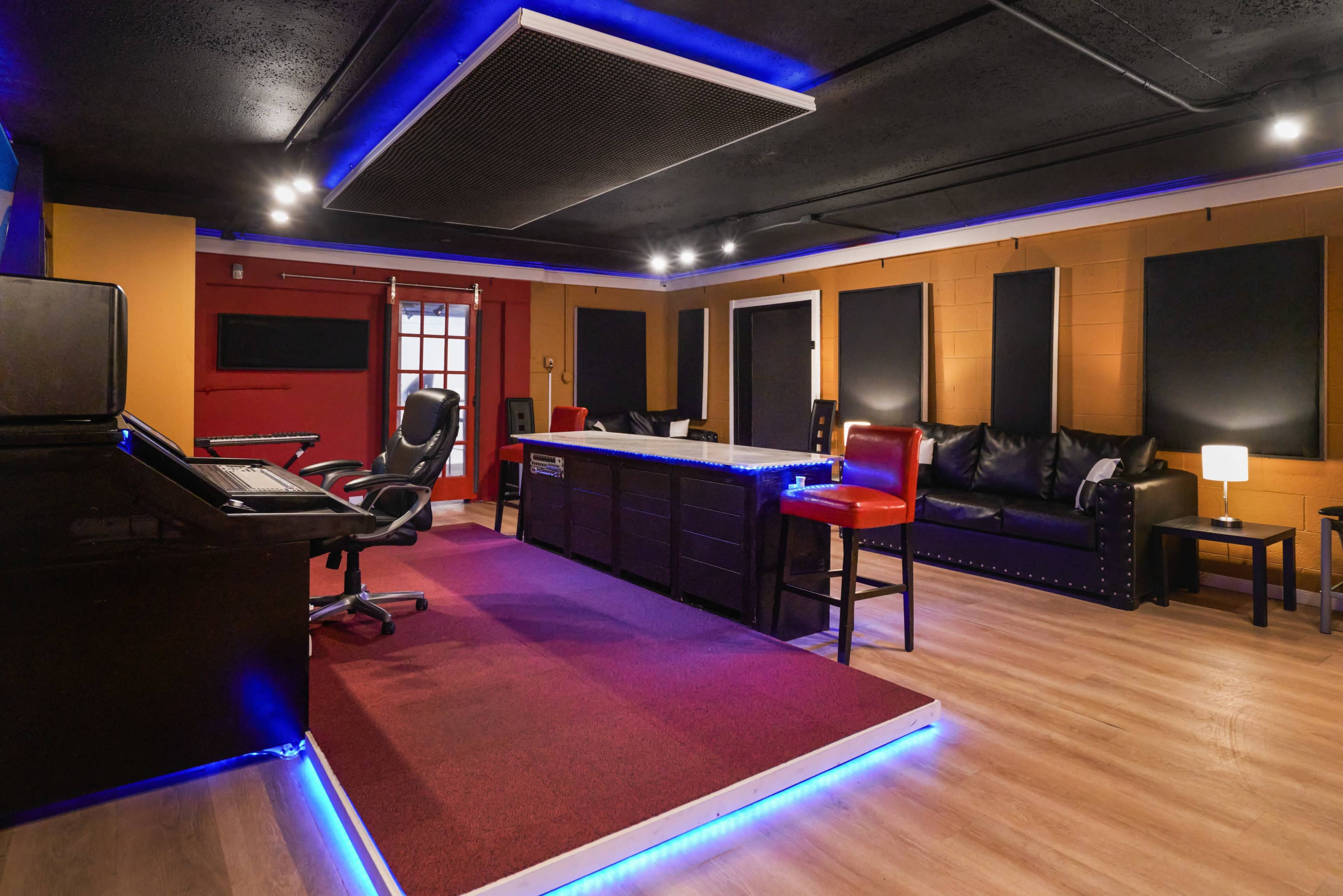 The image shows a recording studio with a mixing desk, black leather seating, and colorful walls illuminated by LED lights.