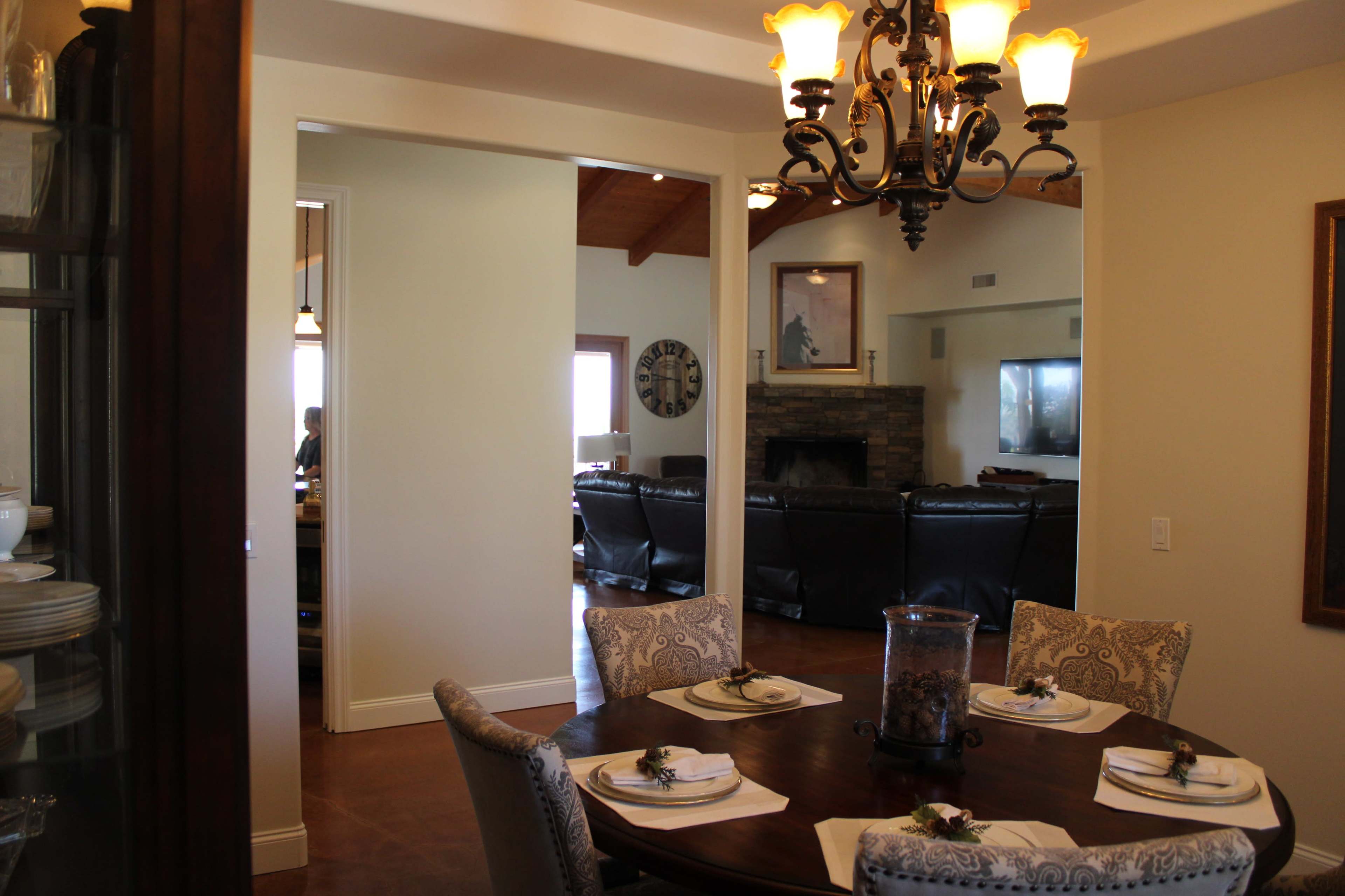 A dining area is visible with a round table set for a meal, leading to a living room featuring a stone fireplace and leather seating.
