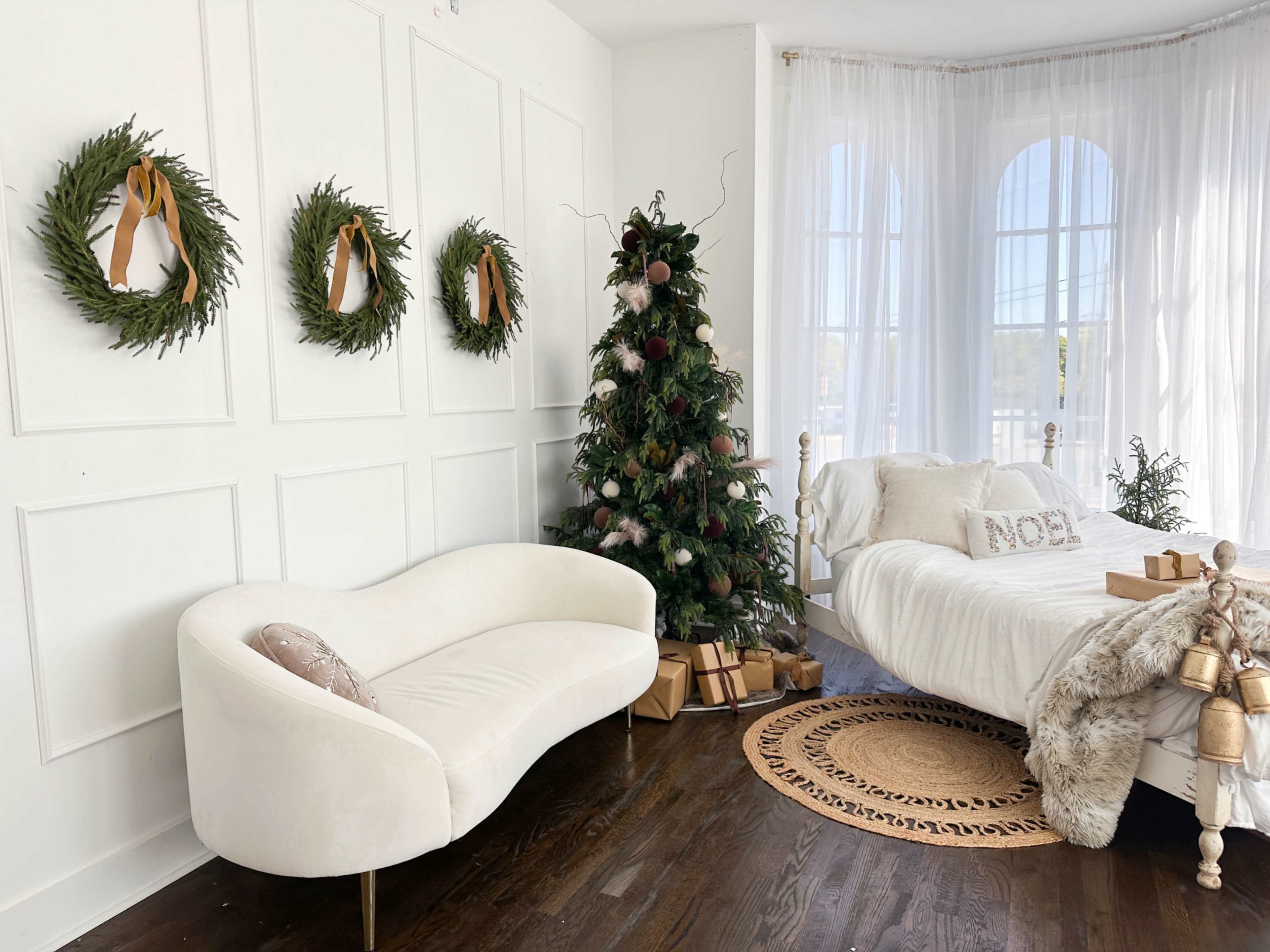 The image shows a cozy bedroom with a white bed adorned with decorative pillows, a Christmas tree decorated with ornaments, and three garlands hung on the wall, alongside a beige couch and wrapped gifts on the floor.
