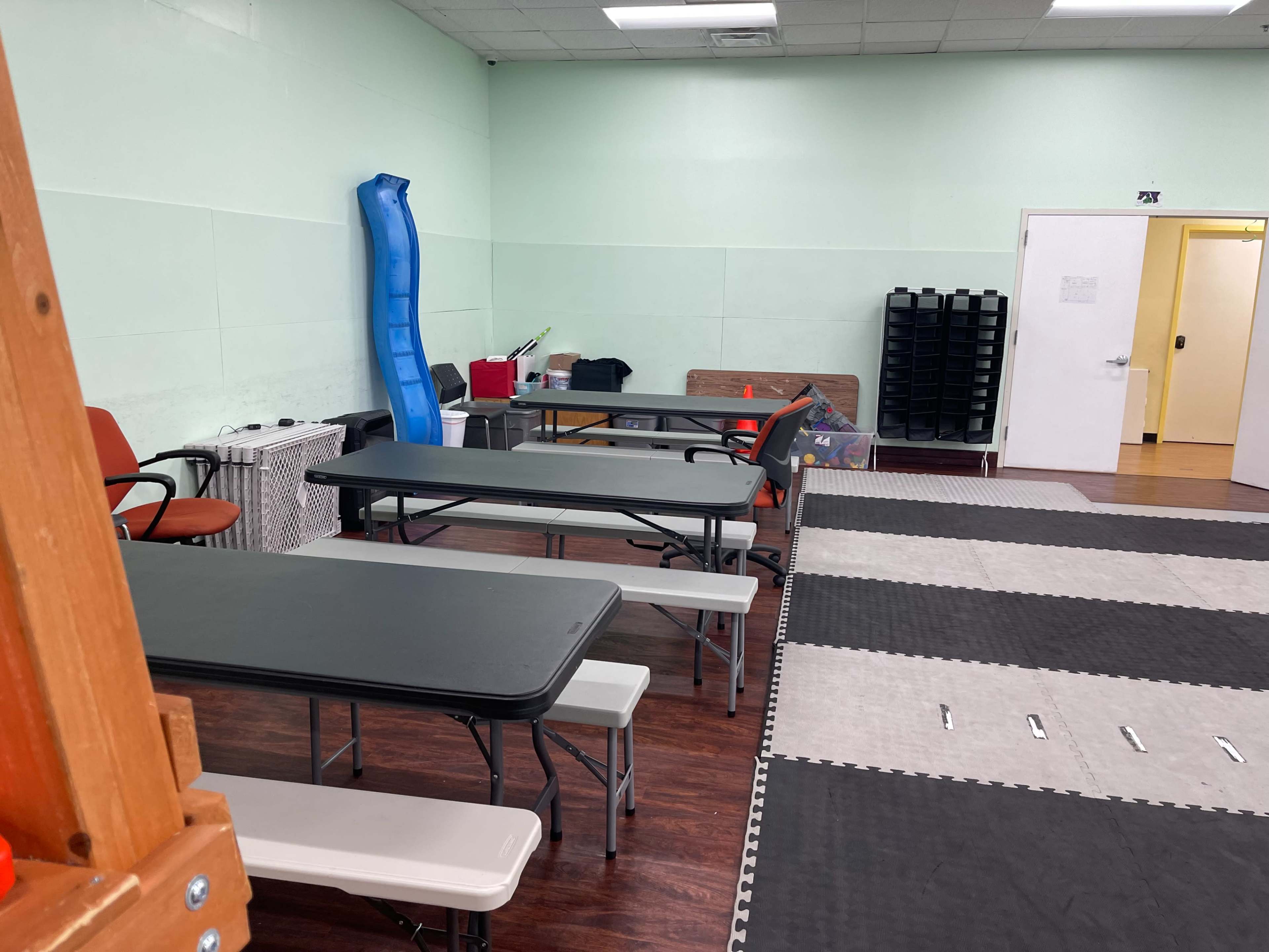 A room with multiple folding tables, chairs, a blue slide, and a mat area on the floor.