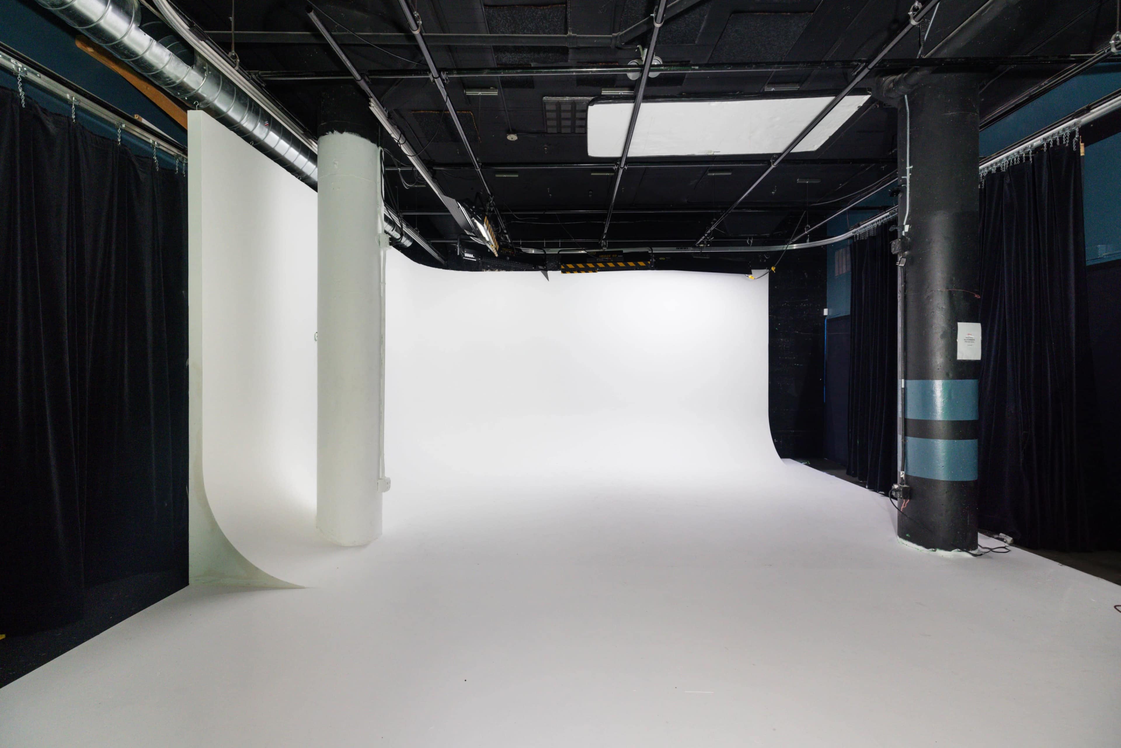 The image shows an empty photography studio with a seamless white backdrop and black curtains along the walls.