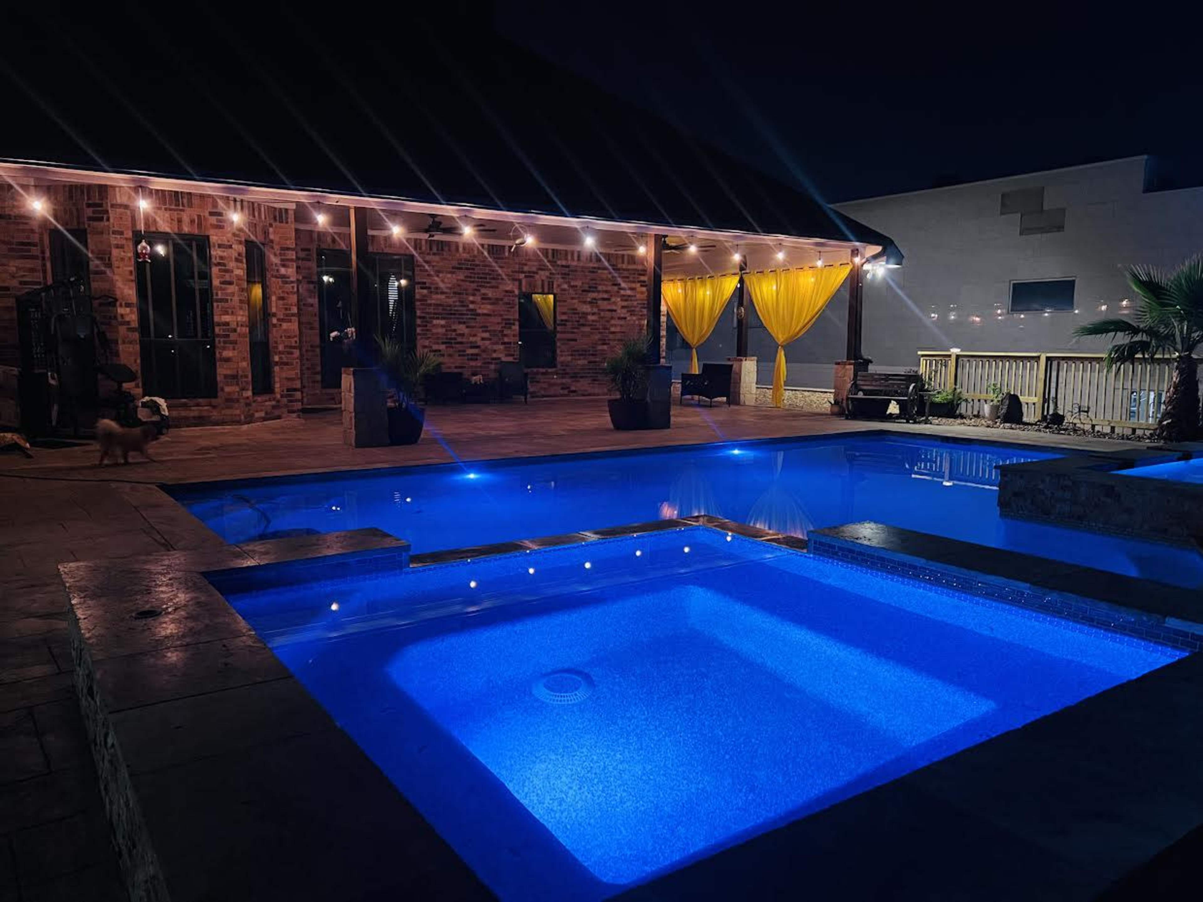 The scene features a well-lit backyard pool area at night with a large pool and a hot tub, surrounded by decorative plants and a brick house in the background.