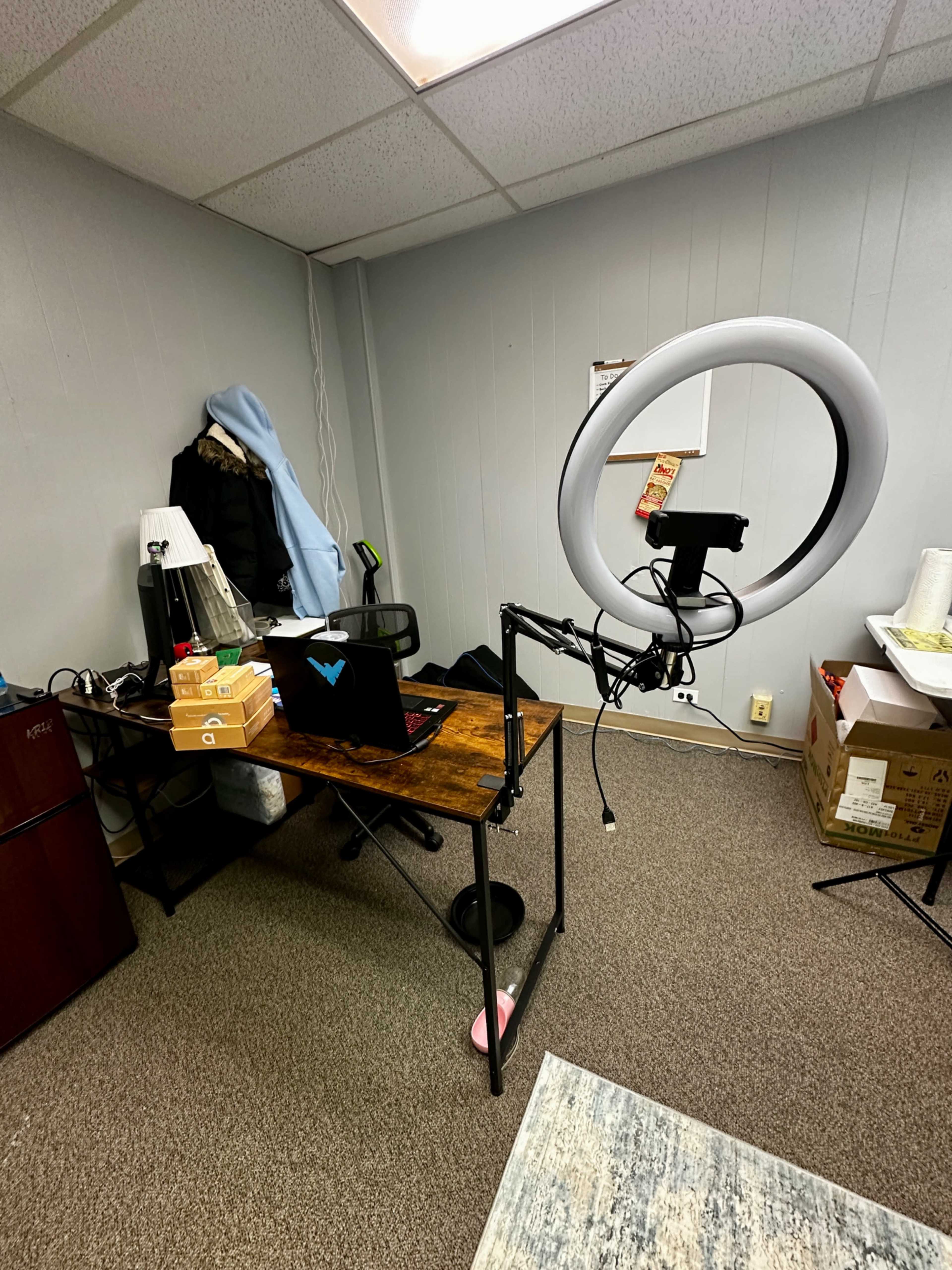 The image shows a small office space with a desk holding a laptop, a ring light mounted on a stand, and boxes stacked nearby.