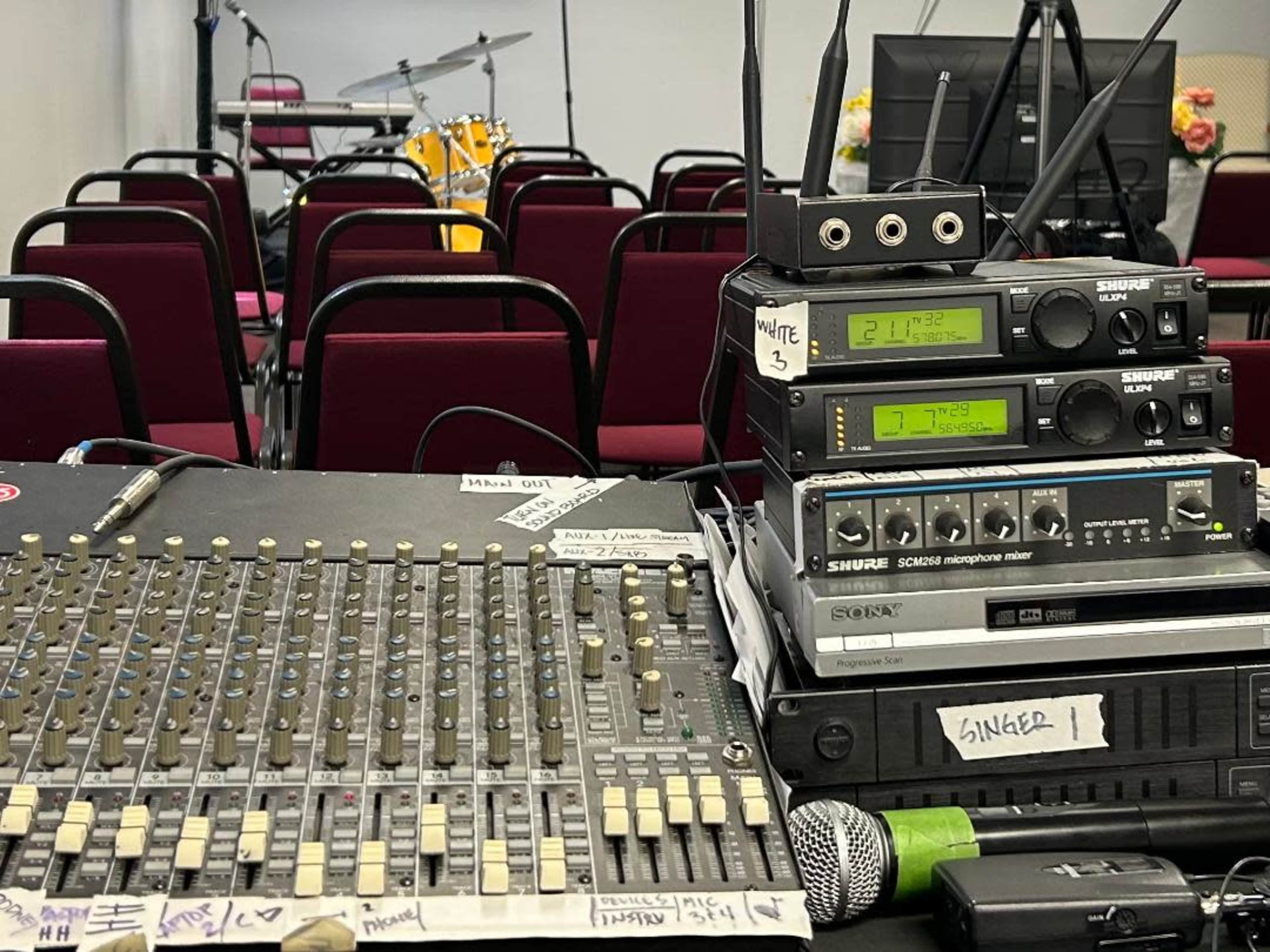 A sound mixing board with several audio devices is positioned in front of empty chairs arranged for a performance, with a drum set visible in the background.