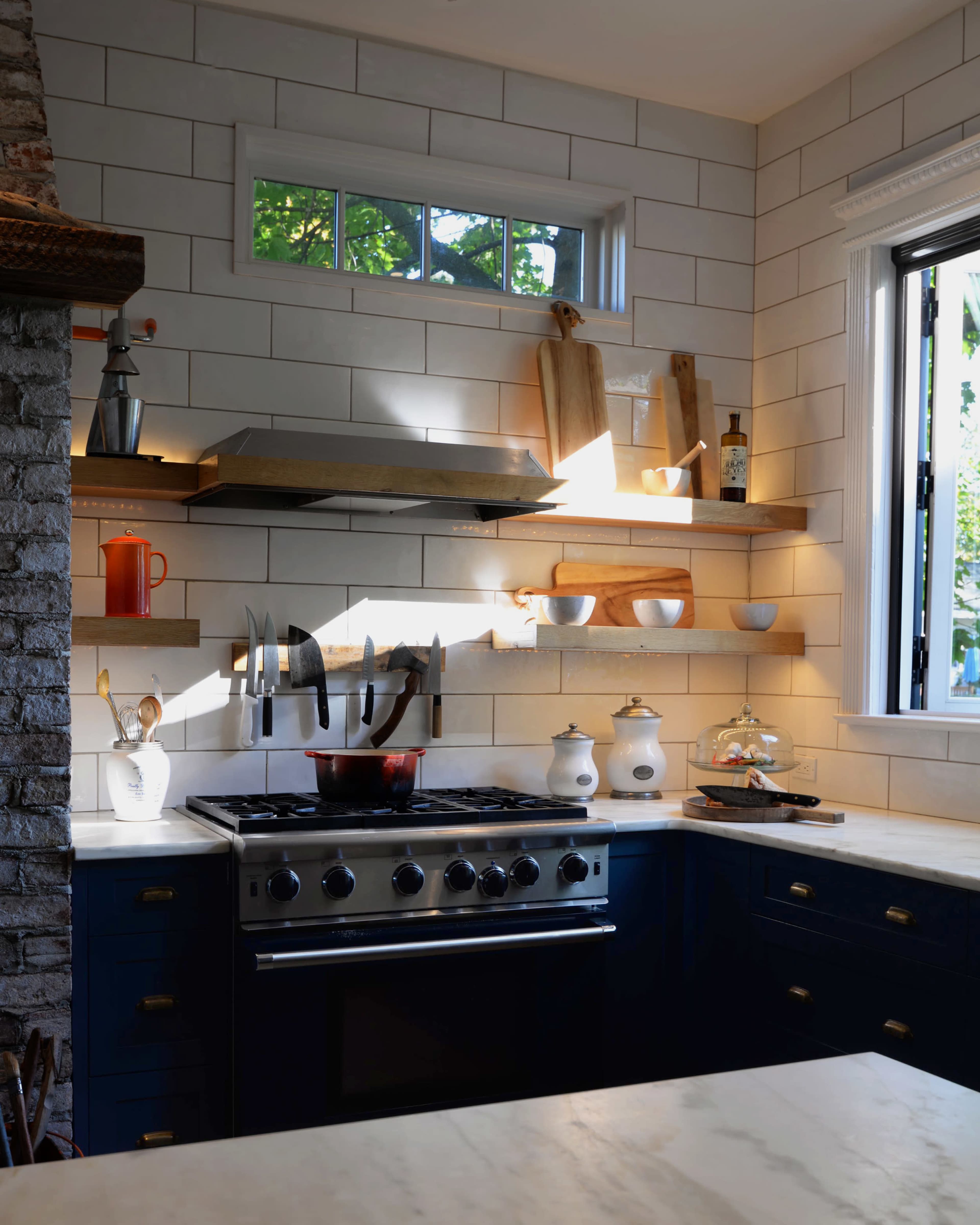 A modern kitchen features a stainless steel stove, open shelves with wooden boards and dishes, and natural light streaming through a window.