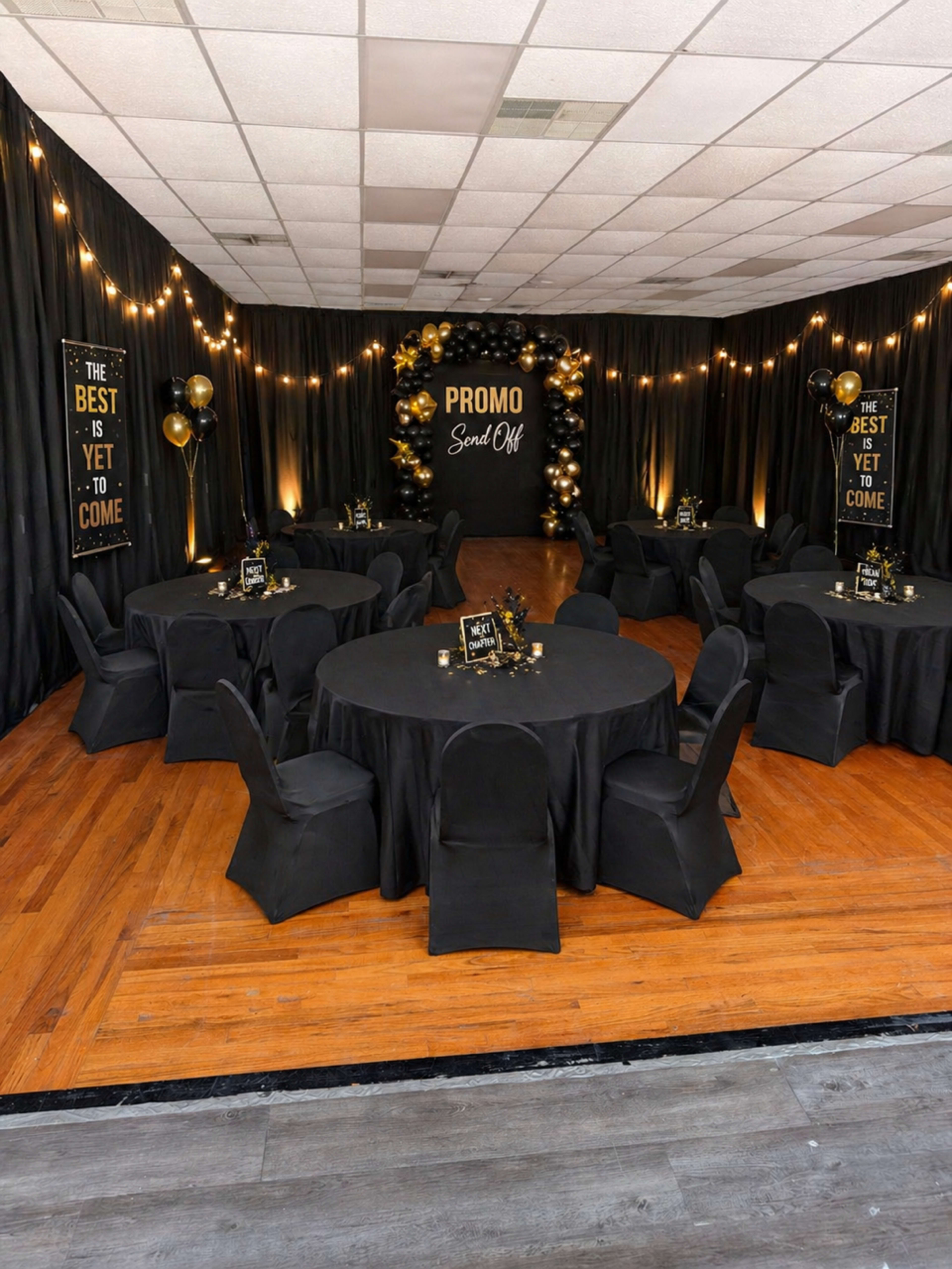 The image shows a decorated event space with black tables and chairs, string lights, and gold and black promotional signage on the walls.