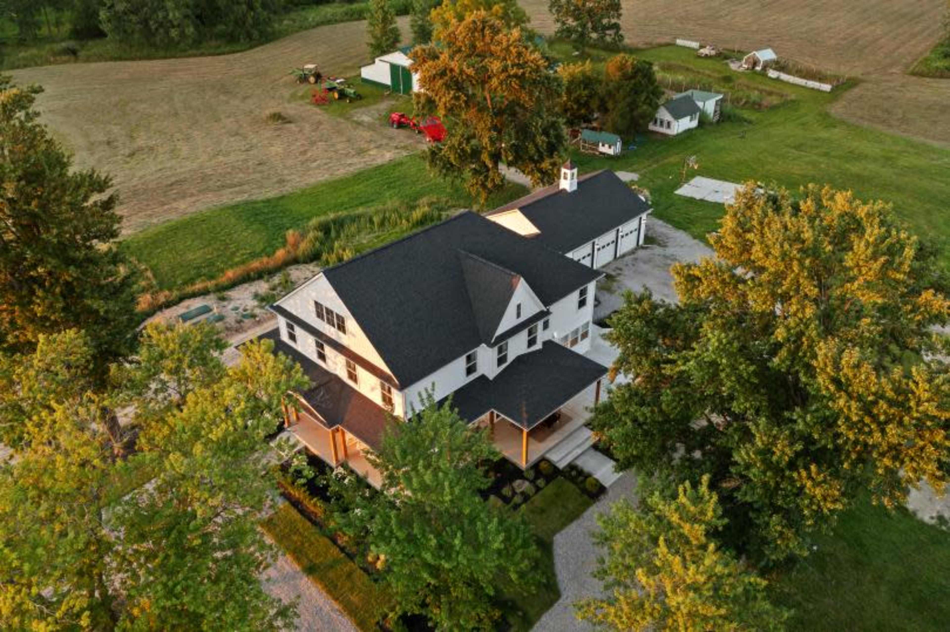 An aerial view shows a large white house with a dark roof surrounded by trees and farmland.