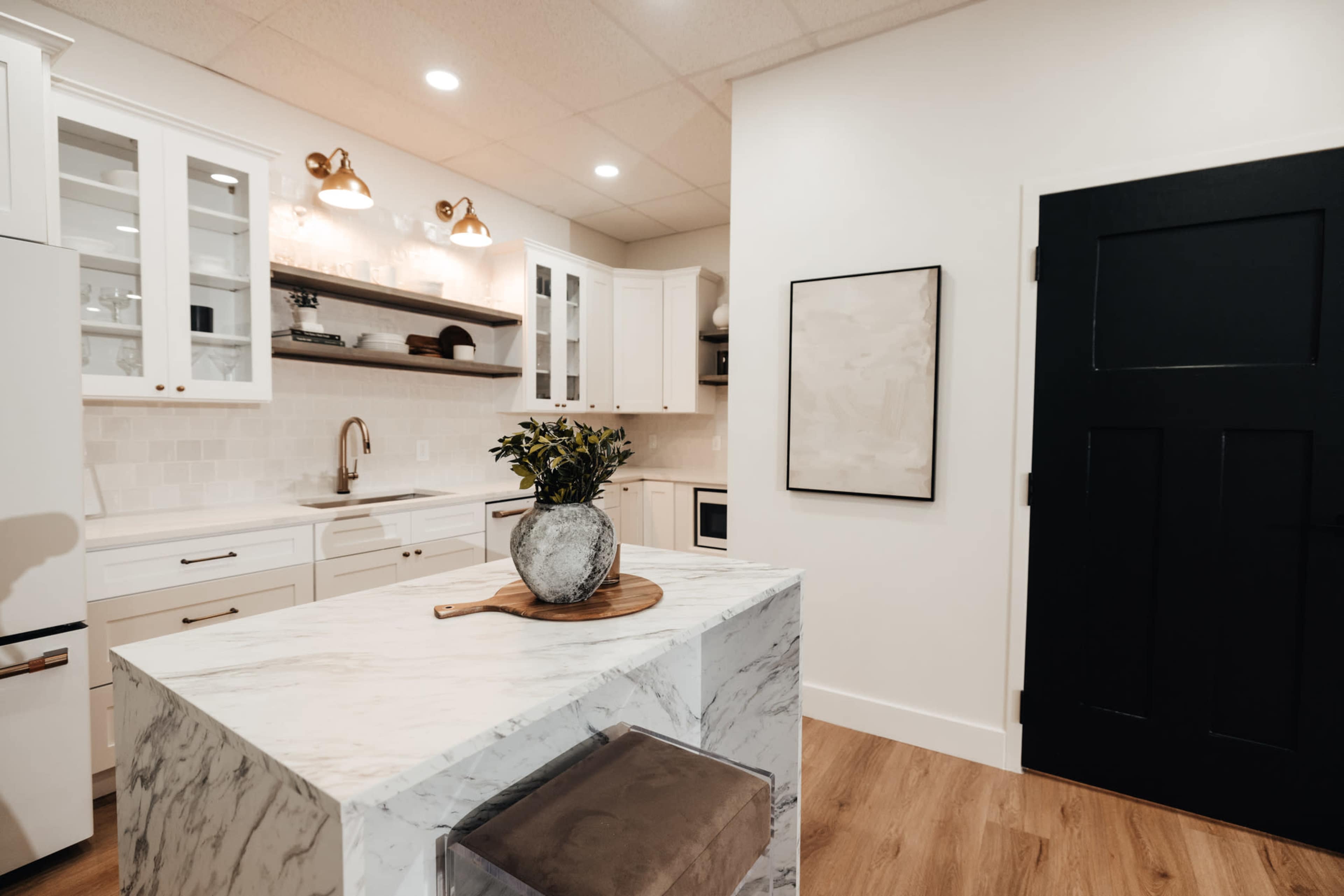 A modern kitchen features white cabinetry, a marble island, and a black door, with warm lighting from wall-mounted fixtures.