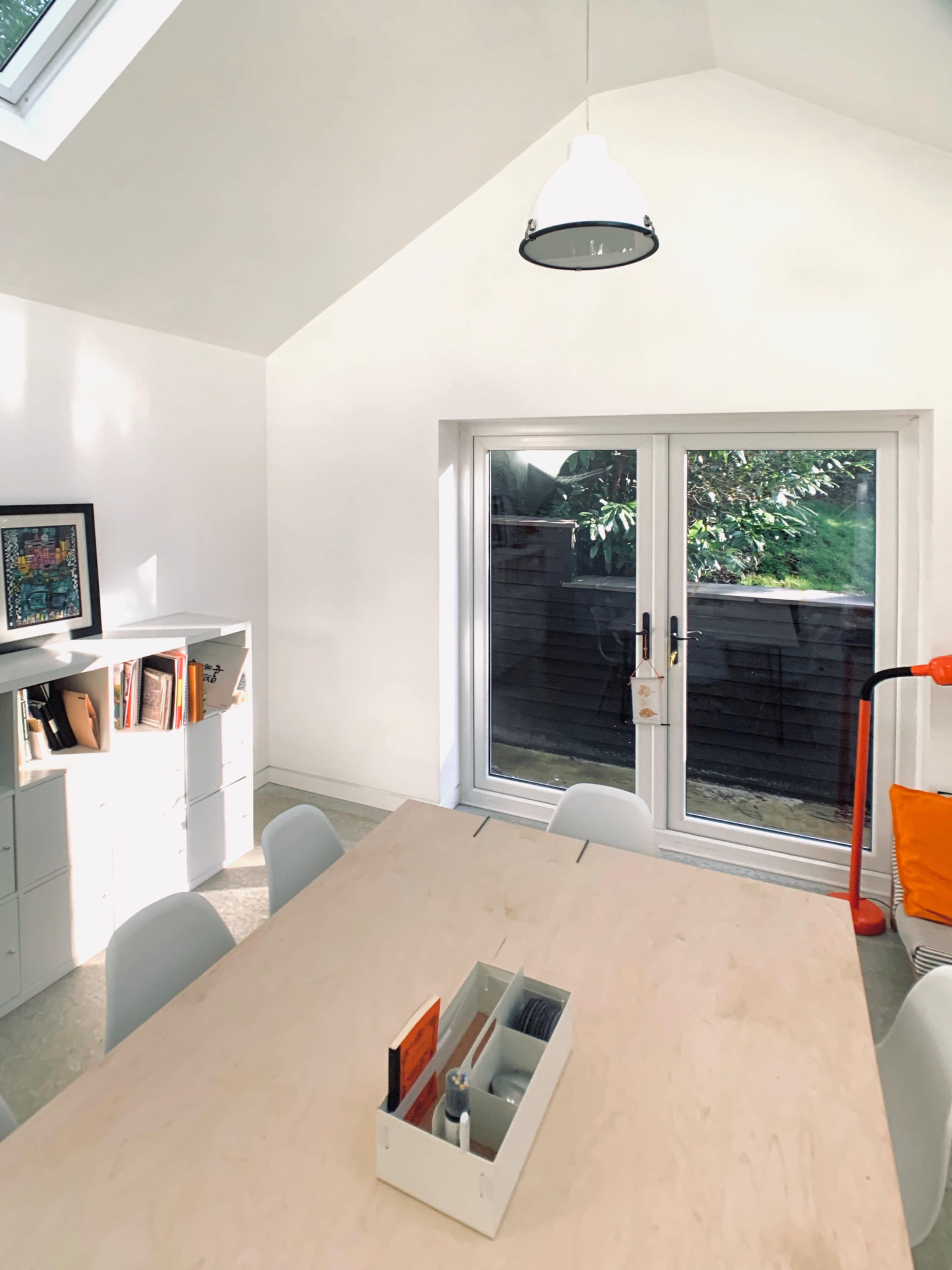 A modern, well-lit dining area features a wooden table surrounded by white chairs, with a bookshelf and large glass doors leading to an outdoor space.