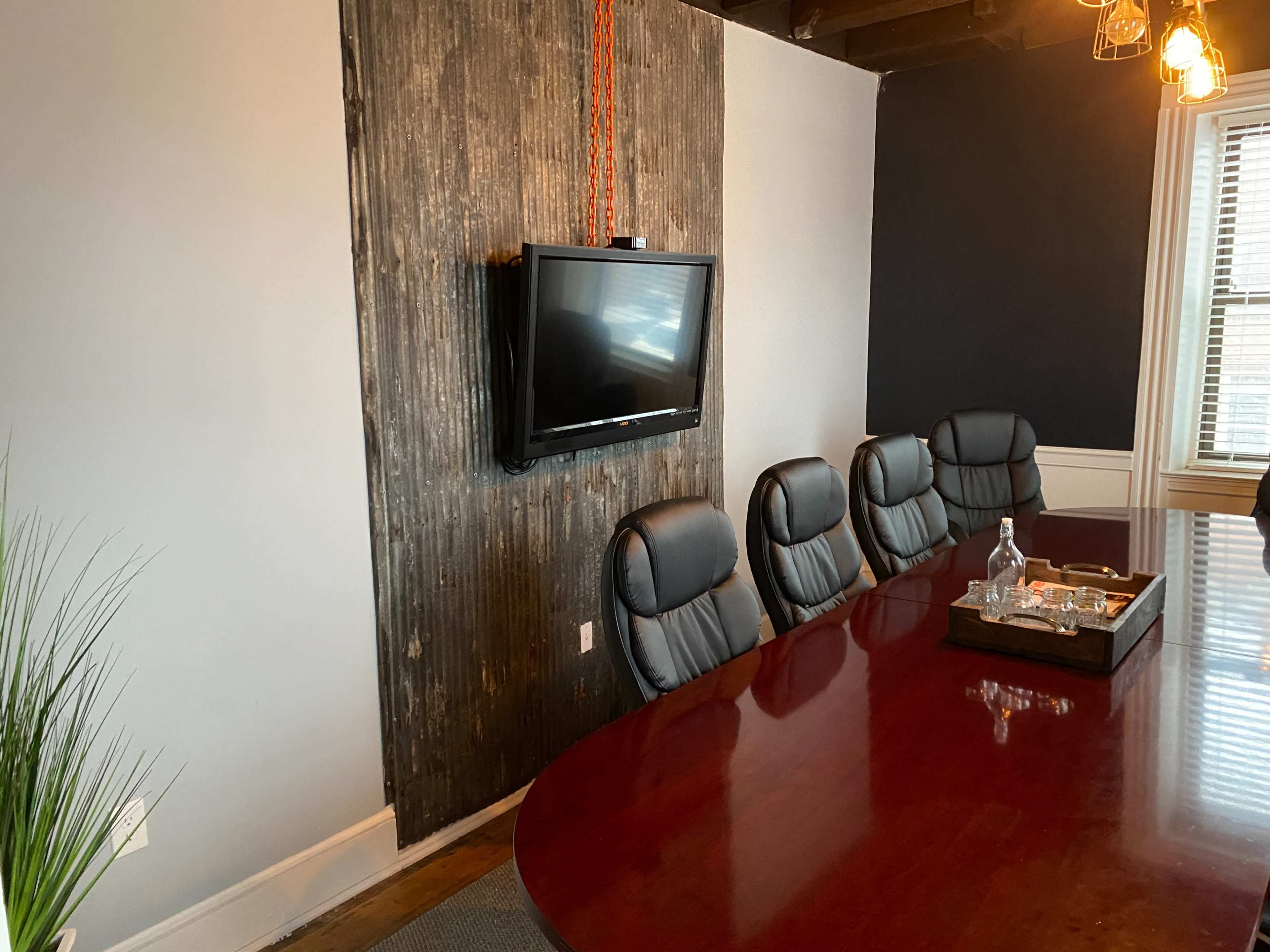A large conference room features a long wooden table with black leather chairs and a wall-mounted television above a textured panel.